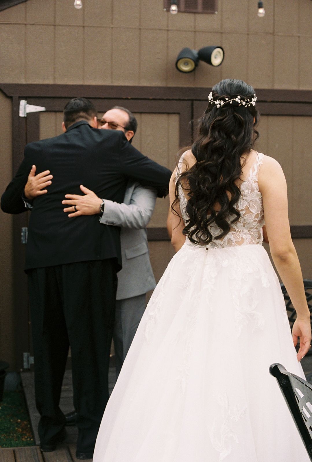 Bride in lace ball gown with floral crown watches groom embrace male guest outside wooden venue.