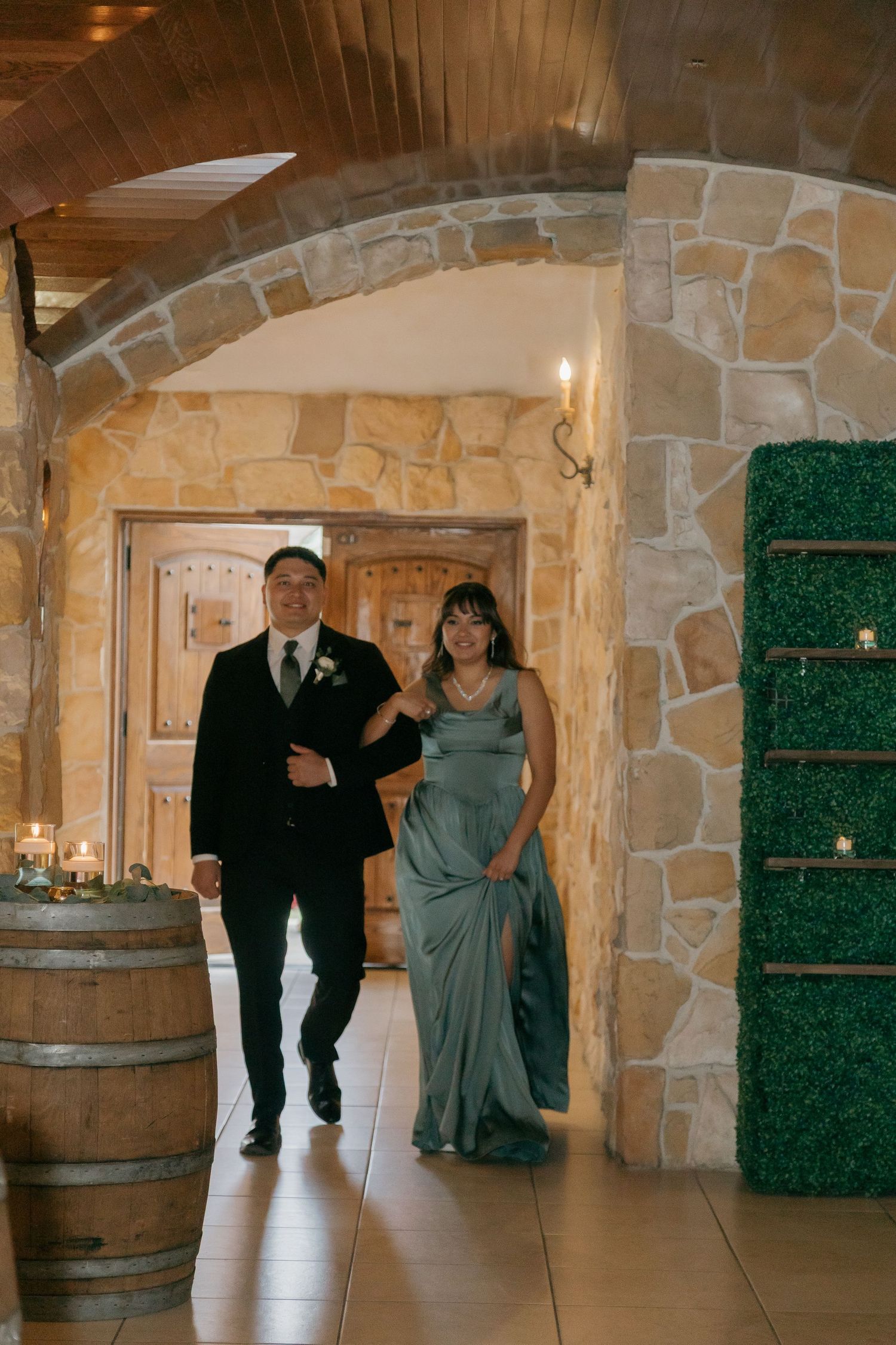 Elegantly dressed couple walks through a stone archway with wine barrel decor at a wedding venue.