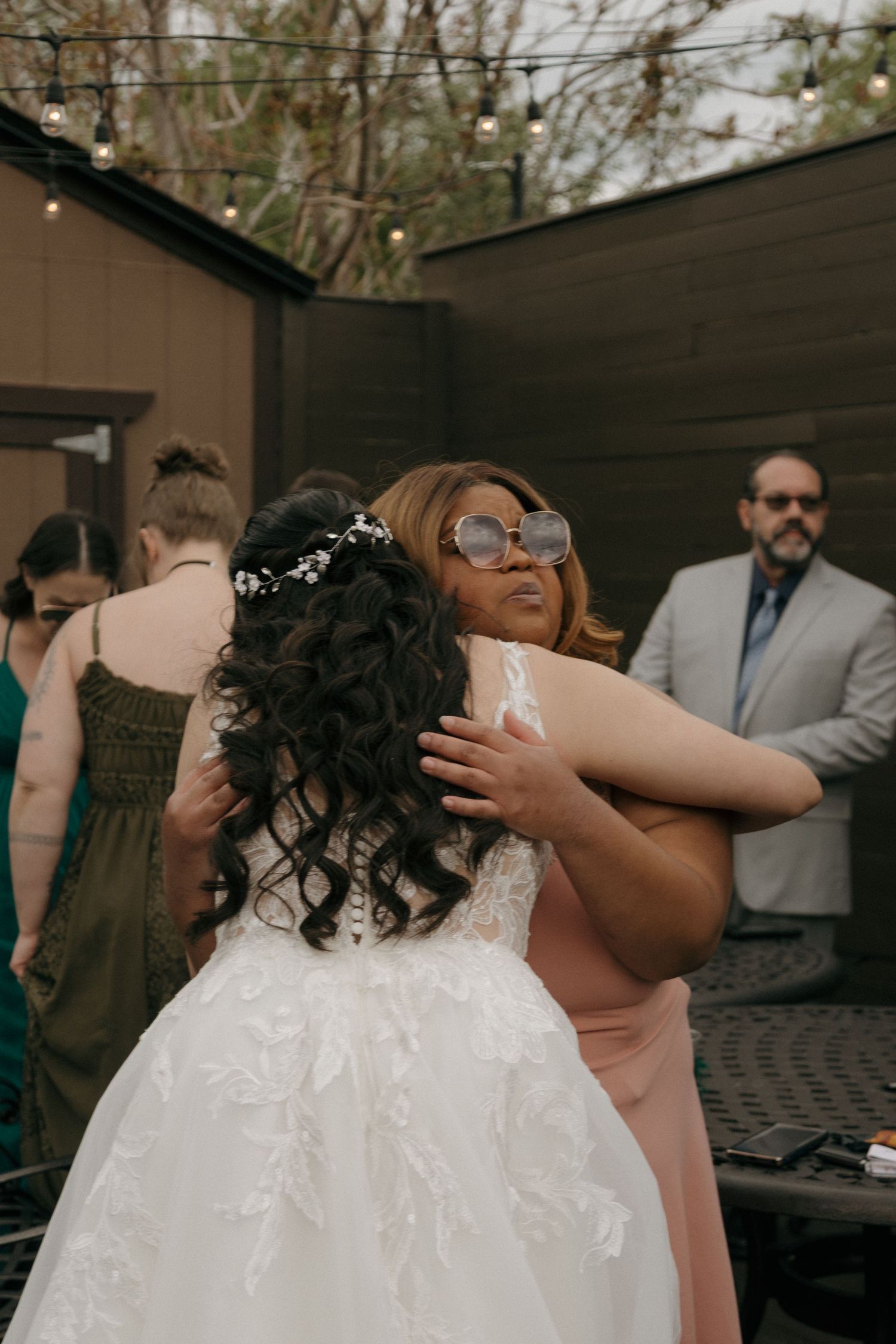 A guest in sunglasses warmly embraces the bride in a lace wedding gown at an outdoor wedding reception.