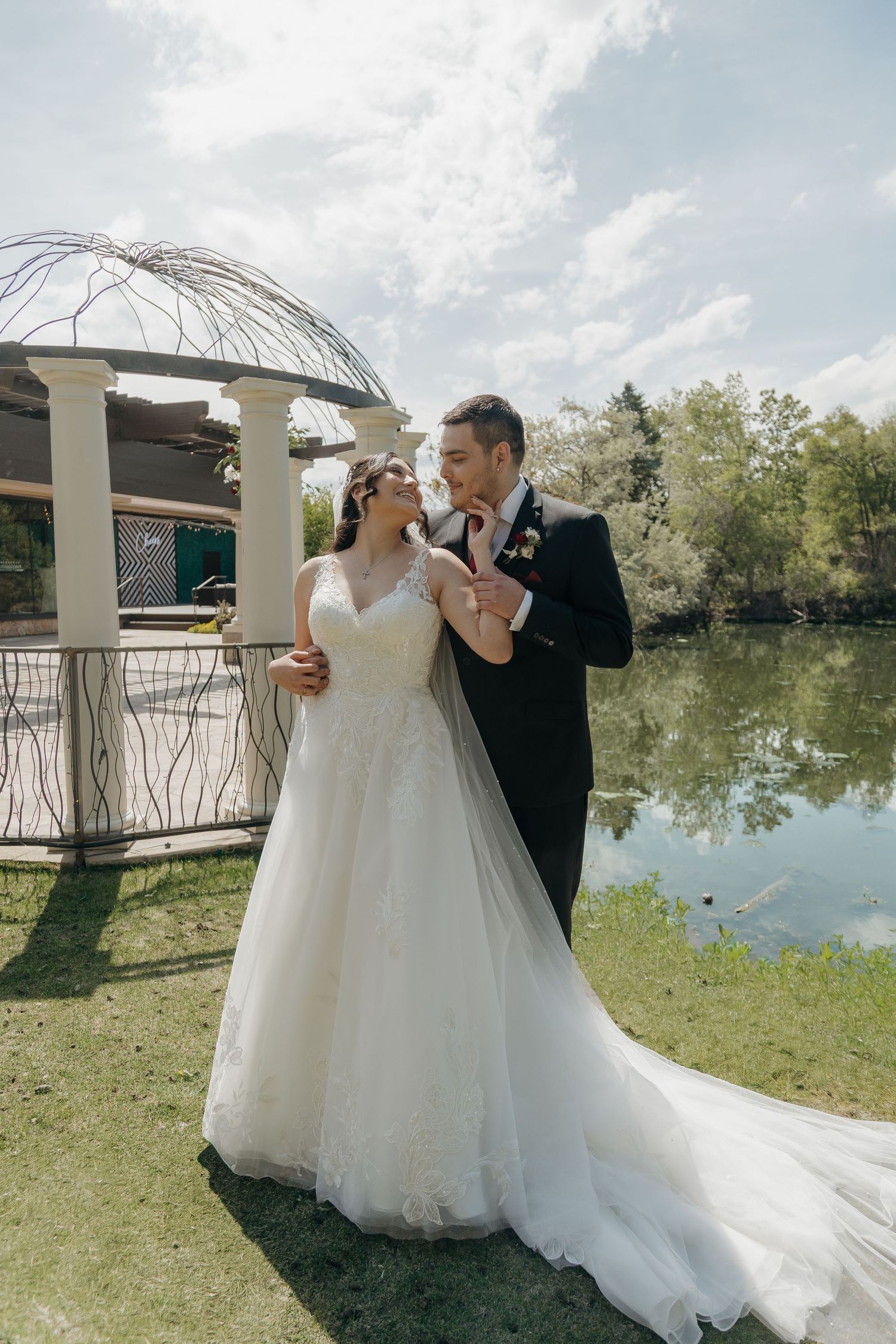 Bride and groom laughing together near a decorative gazebo and pond on a sunny wedding day.