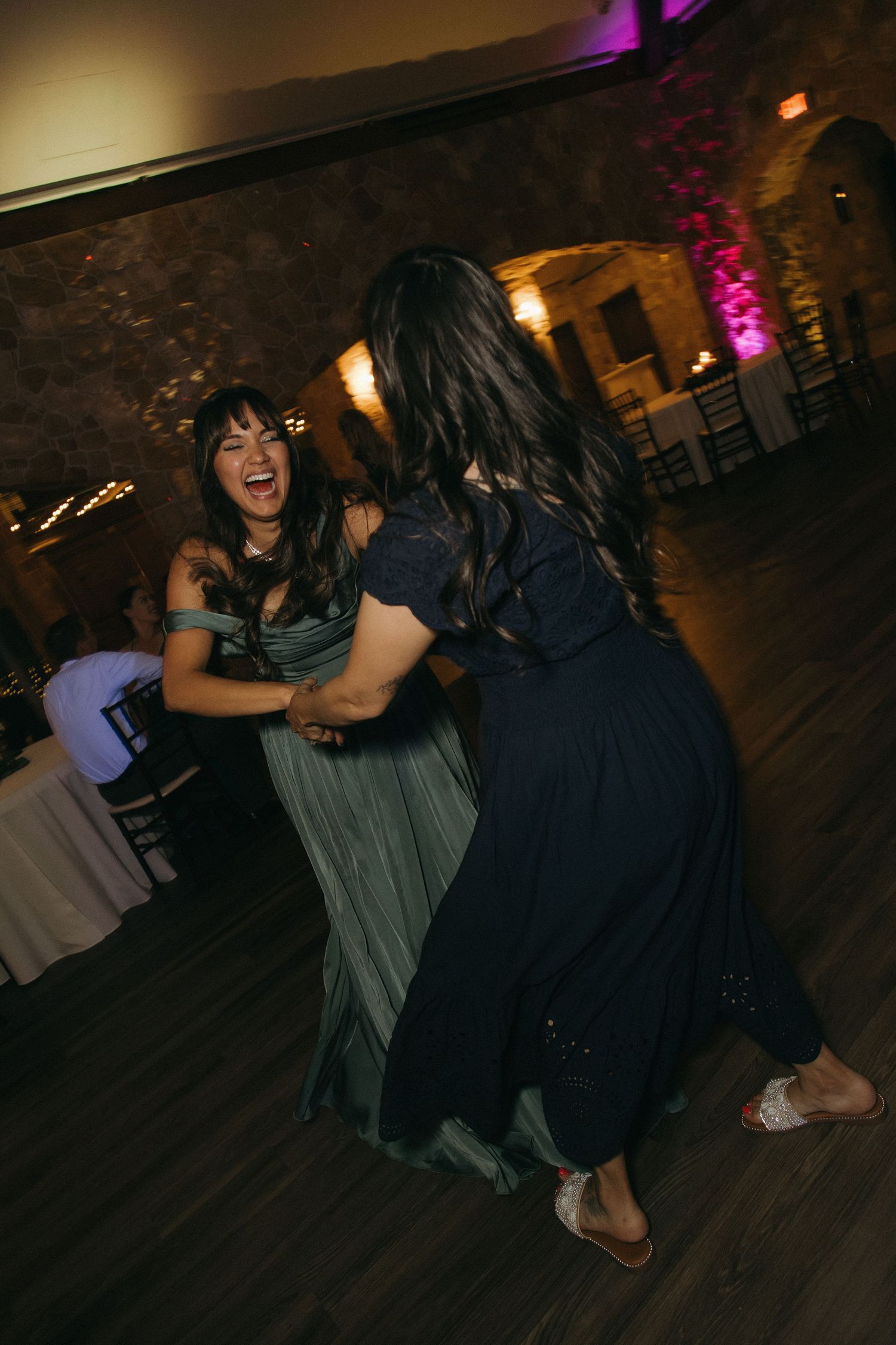 Two women in formal dresses laugh and dance together at dimly lit wedding reception.
