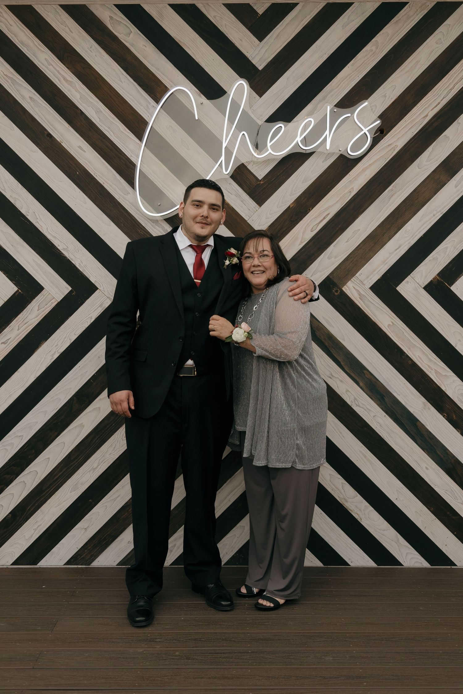 Man in black suit and woman in gray outfit pose together in front of chevron Cheers backdrop.