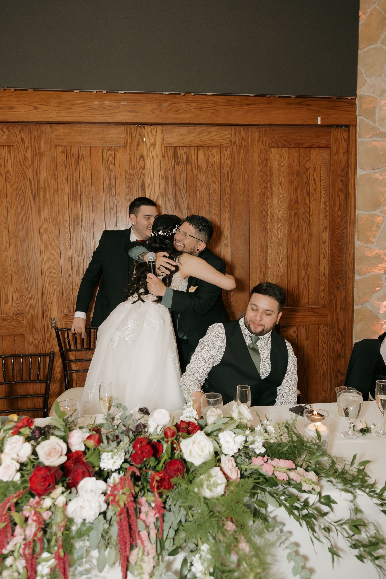 Groomsman hugging bride at the wedding reception head table with red and white florals.