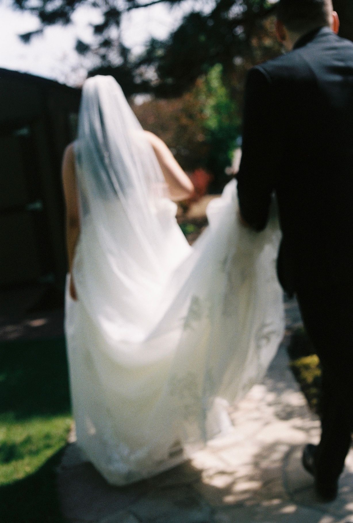 Bride in flowing white gown and veil walking with groom on stone path in outdoor garden setting.