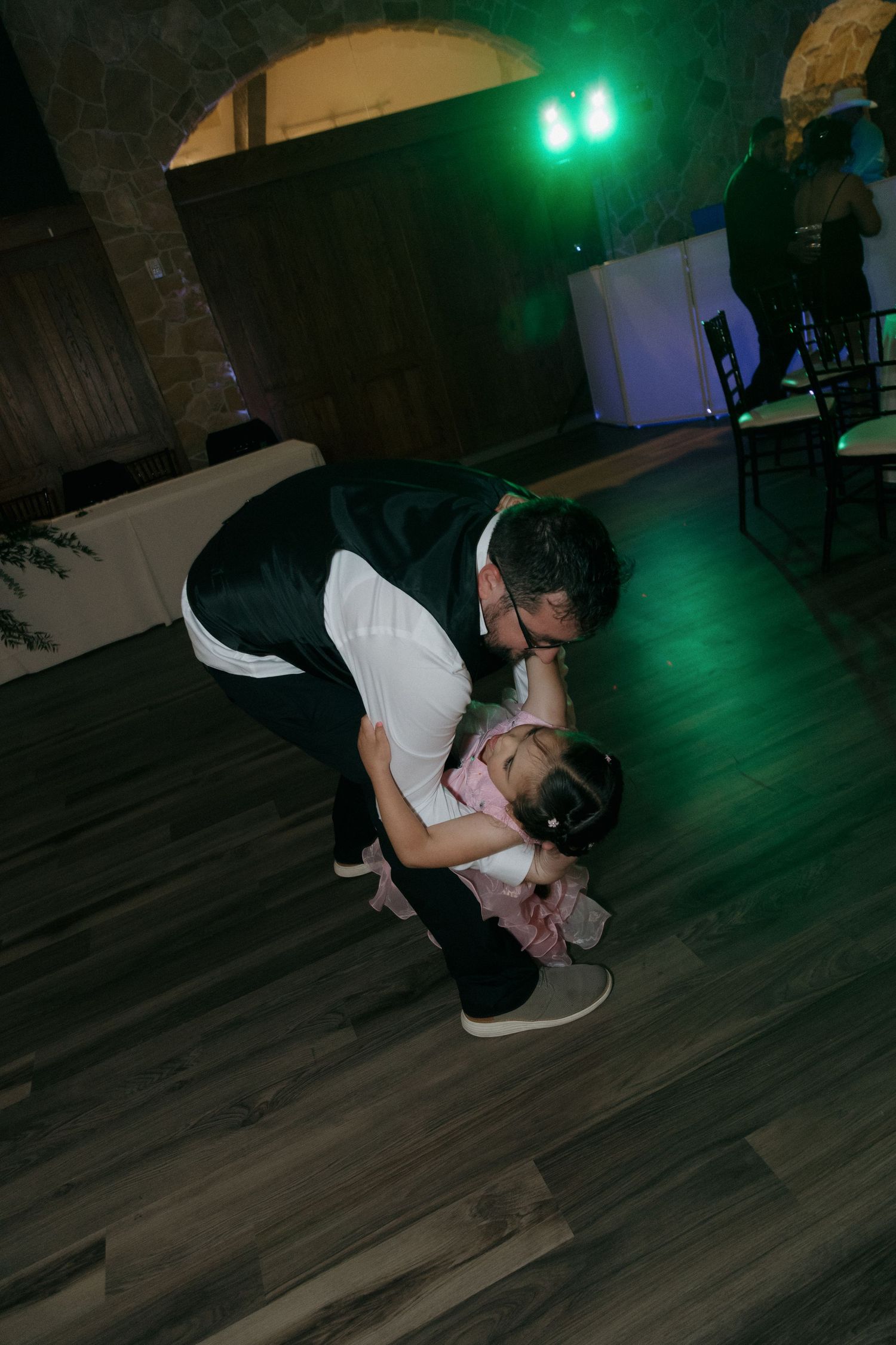 Groom tenderly dips and kisses young flower girl on wedding reception dance floor.