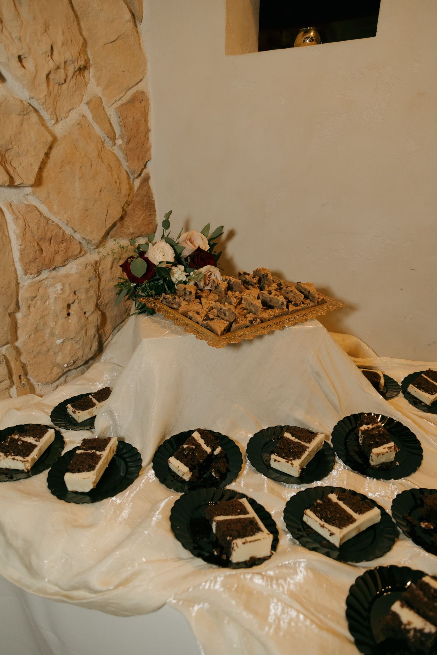 Wedding dessert table featuring sliced cake on black plates and cookies near a stone wall.