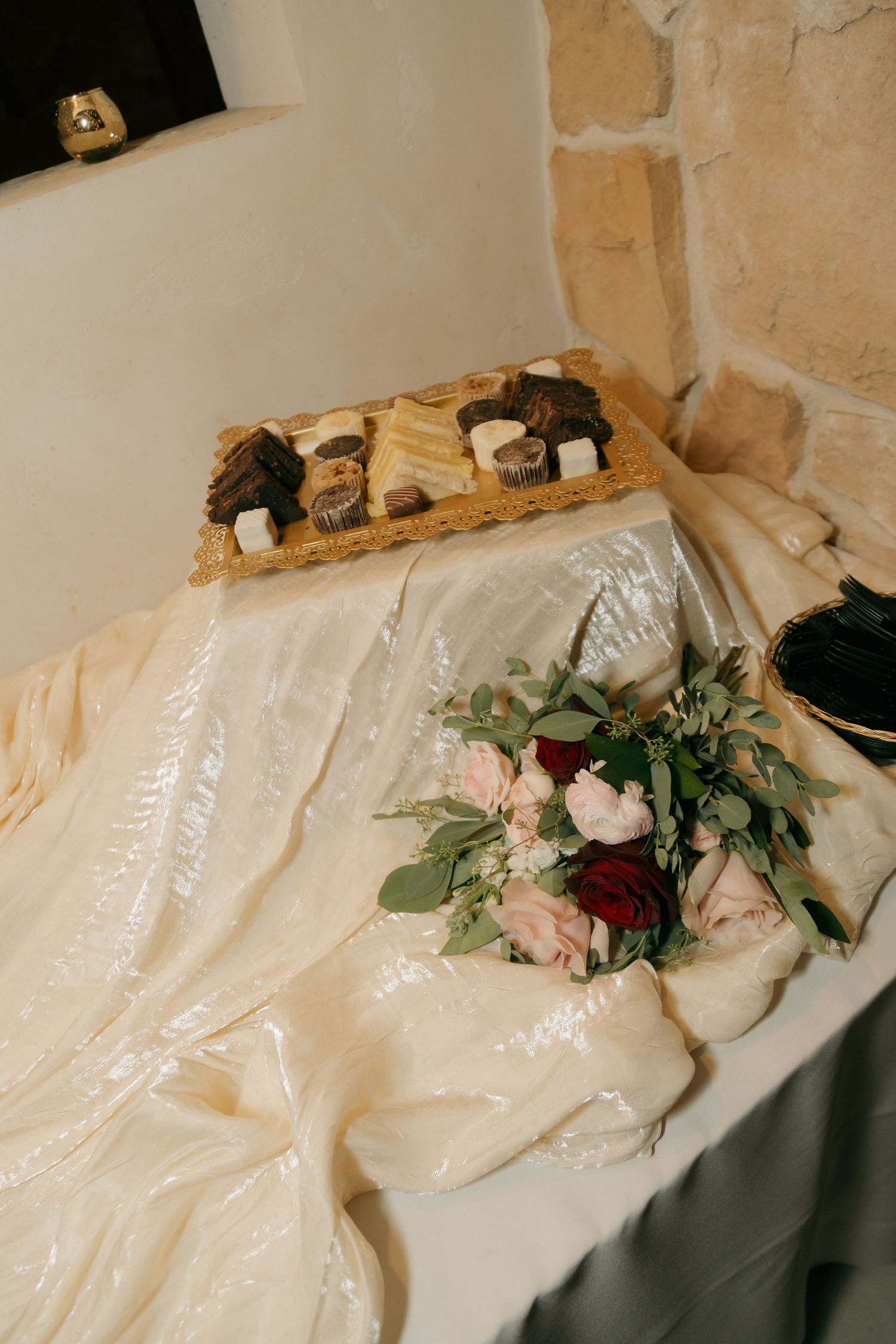 Bridal bouquet of pink and red roses with greenery rests on white tablecloth near dessert tray.