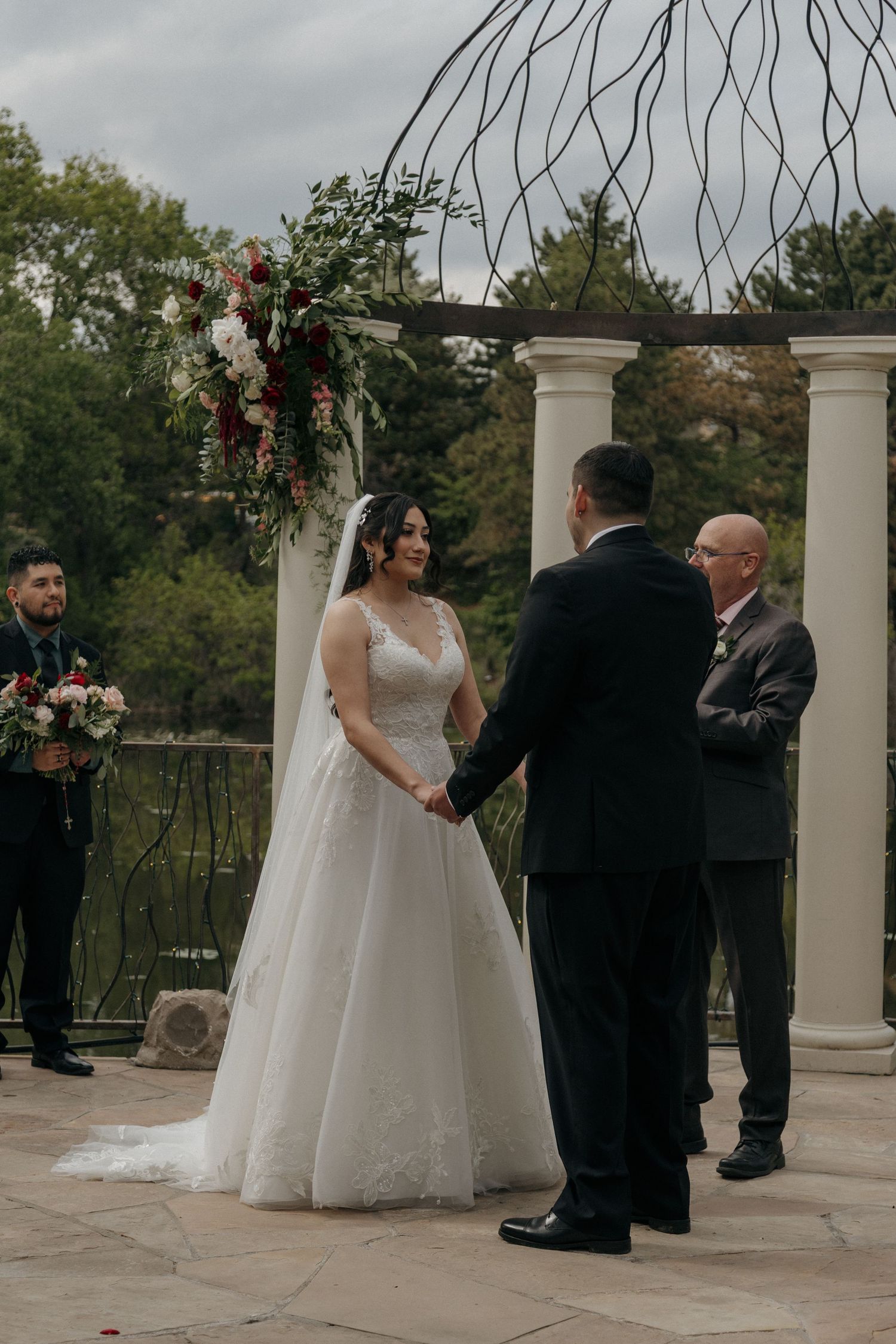 Smiling bride holds grooms hands at outdoor wedding ceremony beneath decorative arch with floral arrangements.
