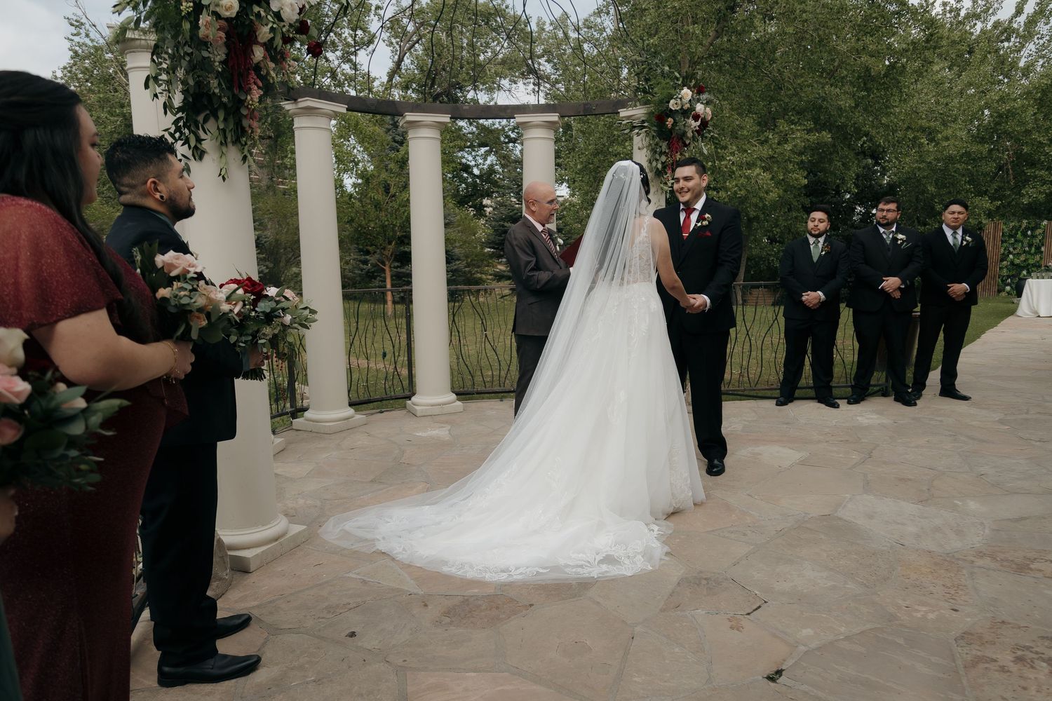 Bride in flowing white gown with cathedral veil walks toward groom at outdoor wedding ceremony with floral arch.