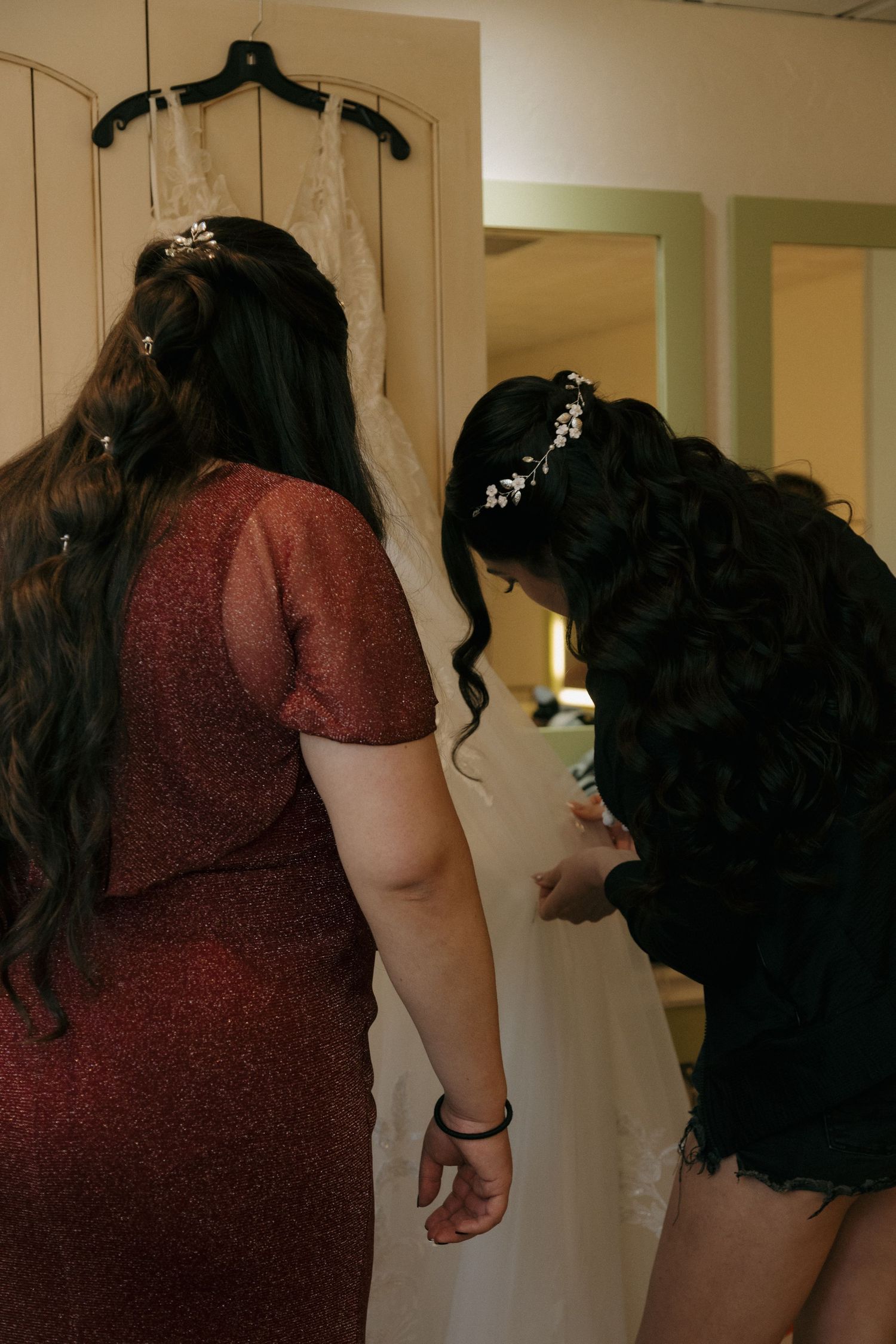 Two dark-haired women in a bridal suite preparing a lace wedding dress hanging on the wall.
