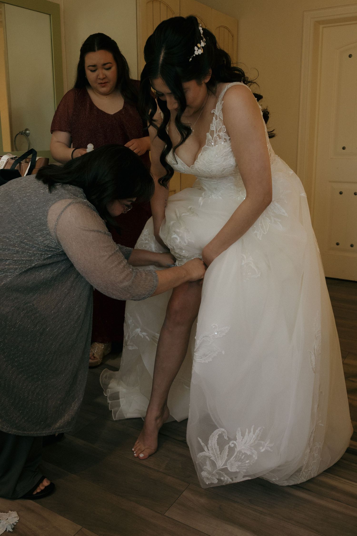 Bride in white lace wedding gown being helped with shoes by two women during bridal preparations.
