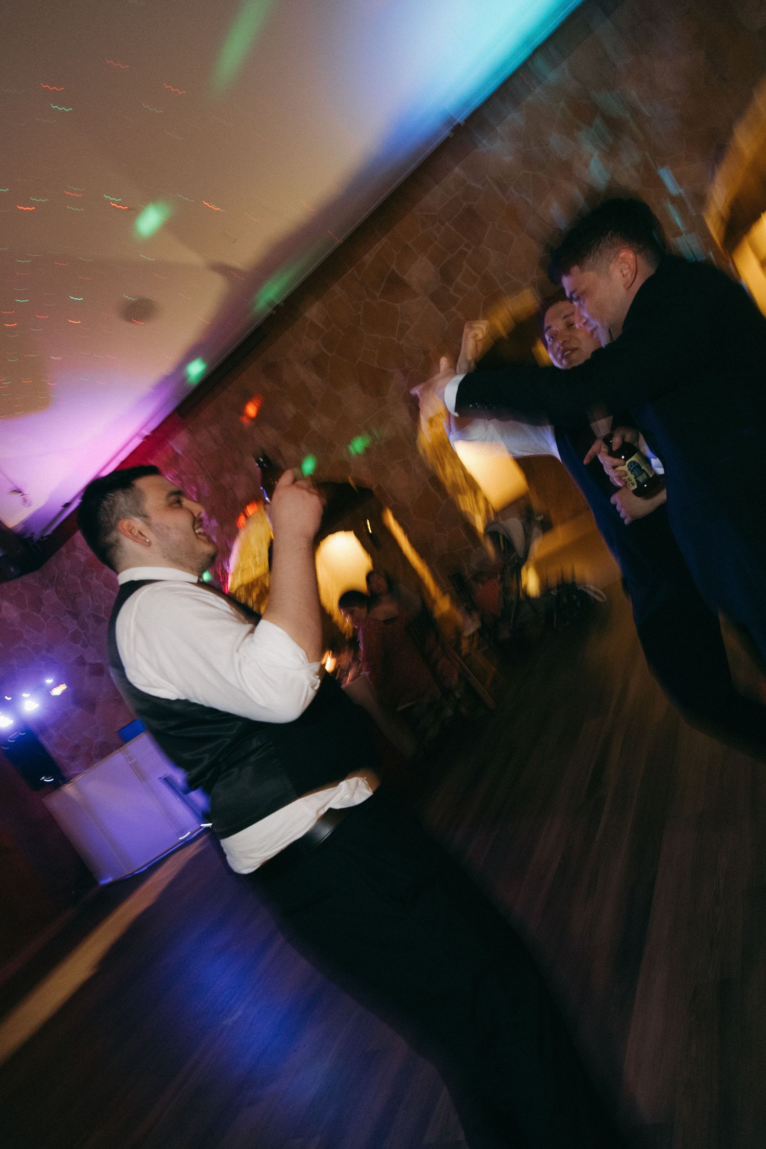 Guests dance energetically on a colorfully lit wedding reception dance floor.