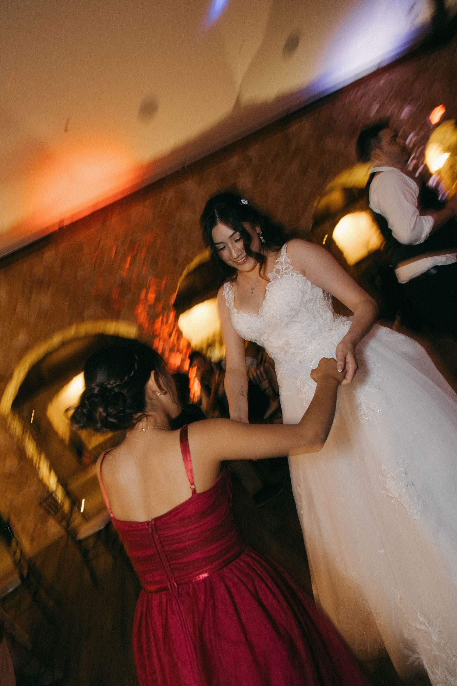 Bride in white lace gown dancing with bridesmaid in red dress at reception.