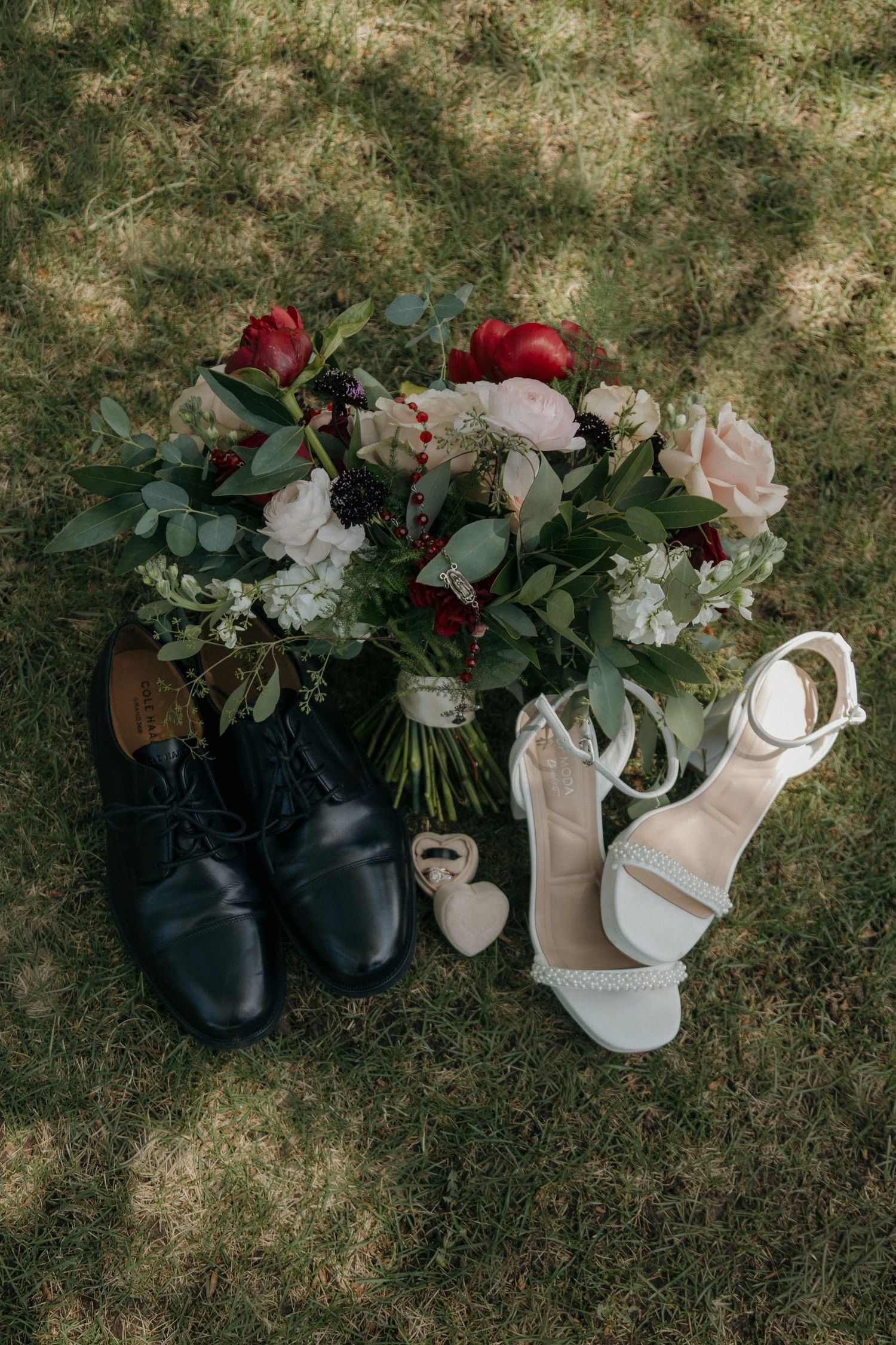 Wedding flat lay with black groom shoes, white bridal heels, and a bouquet of red and pink roses on grass.