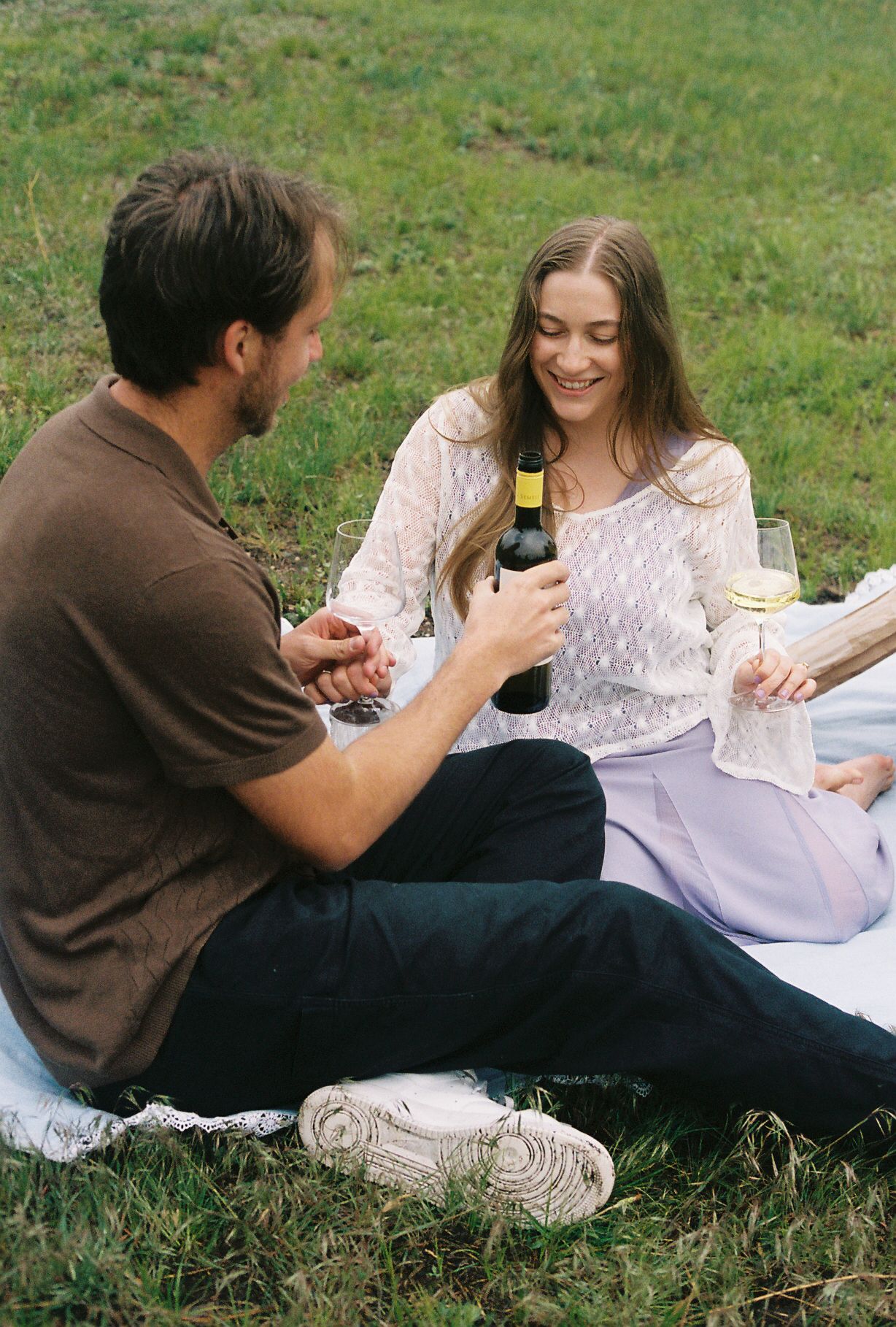 Couple enjoying a picnic on grass, woman in white lace top pouring white wine, man in brown shirt holding glass.