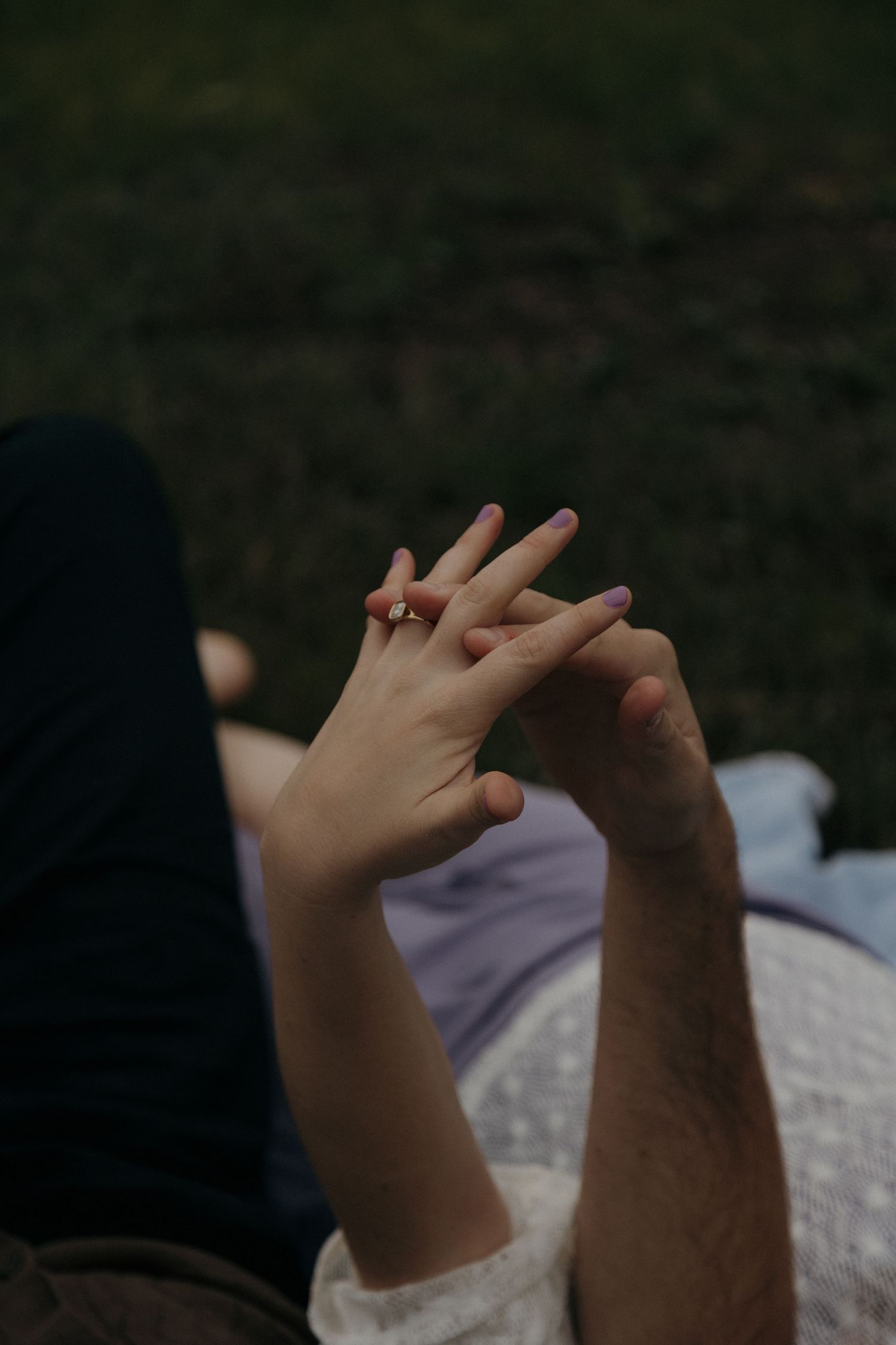 Close-up of couple holding hands with engagement ring on grassy background.