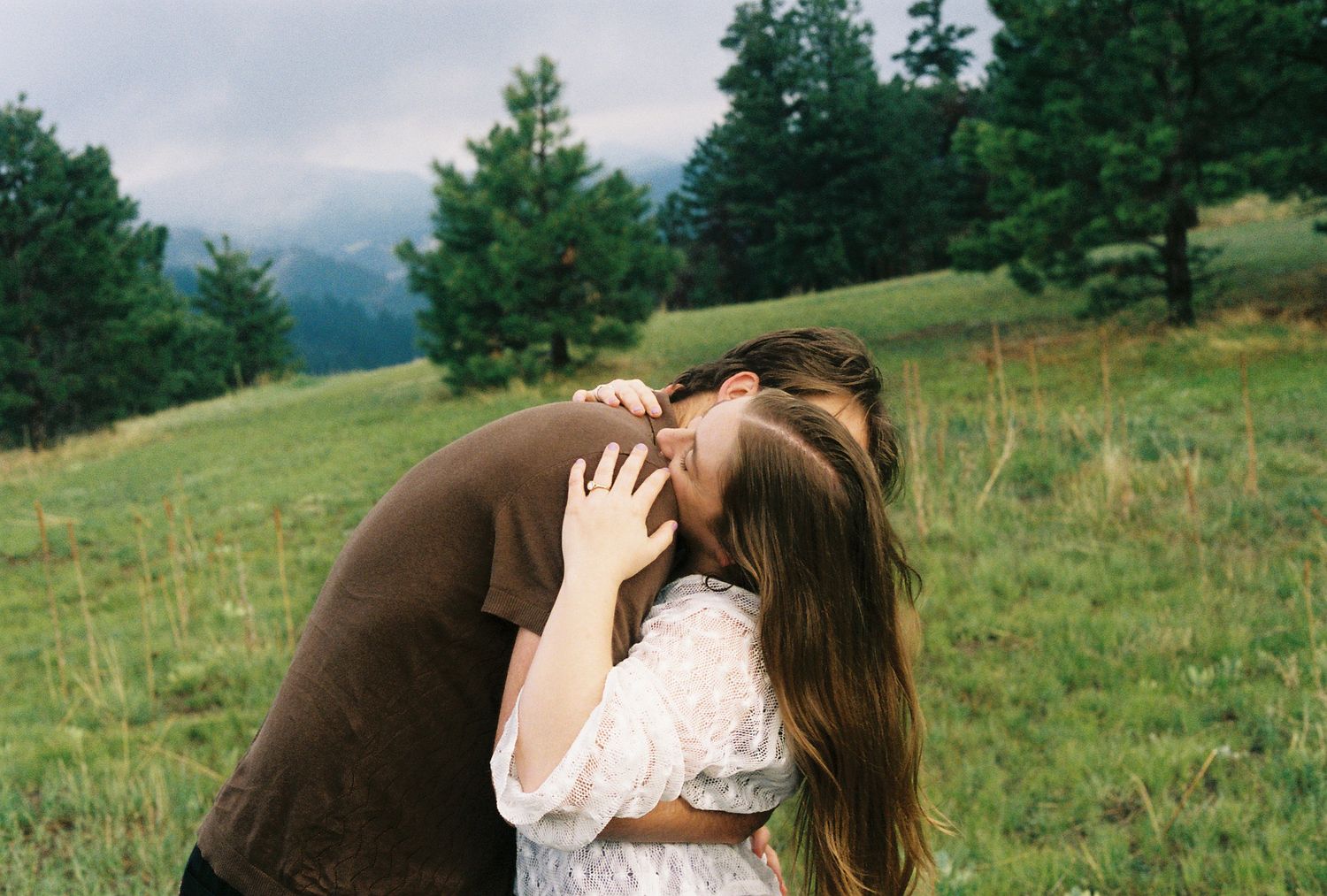 Couple sharing a tender kiss in a green mountain meadow surrounded by pine trees.
