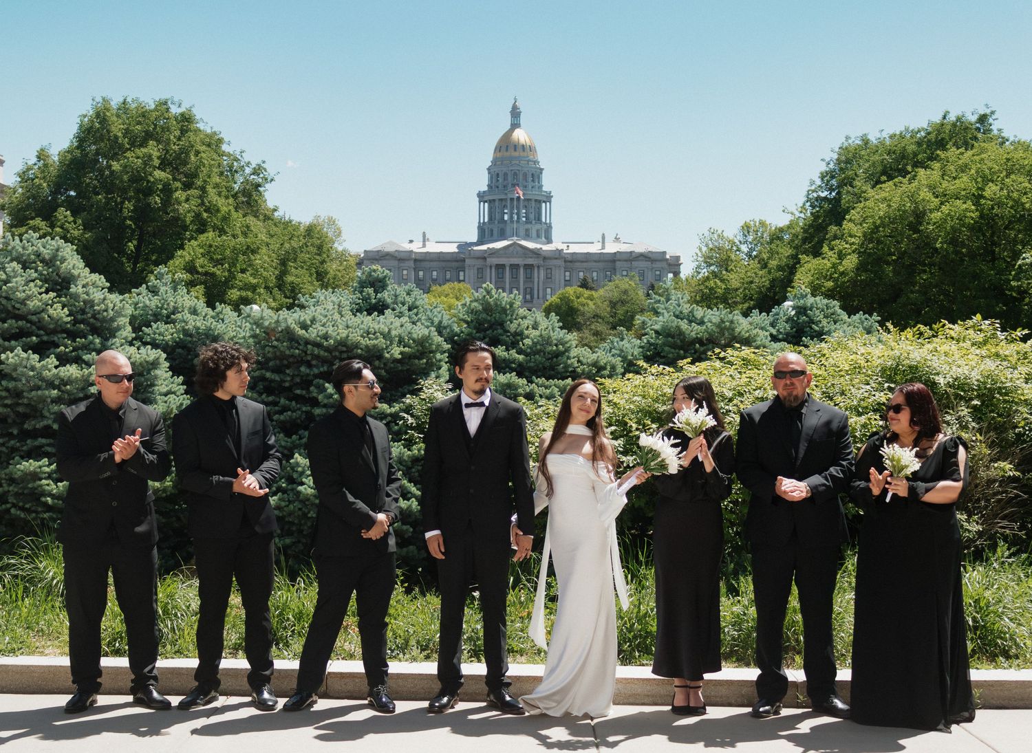 Wedding party members in black attire strike playful poses in a line against a sunny outdoor garden backdrop.