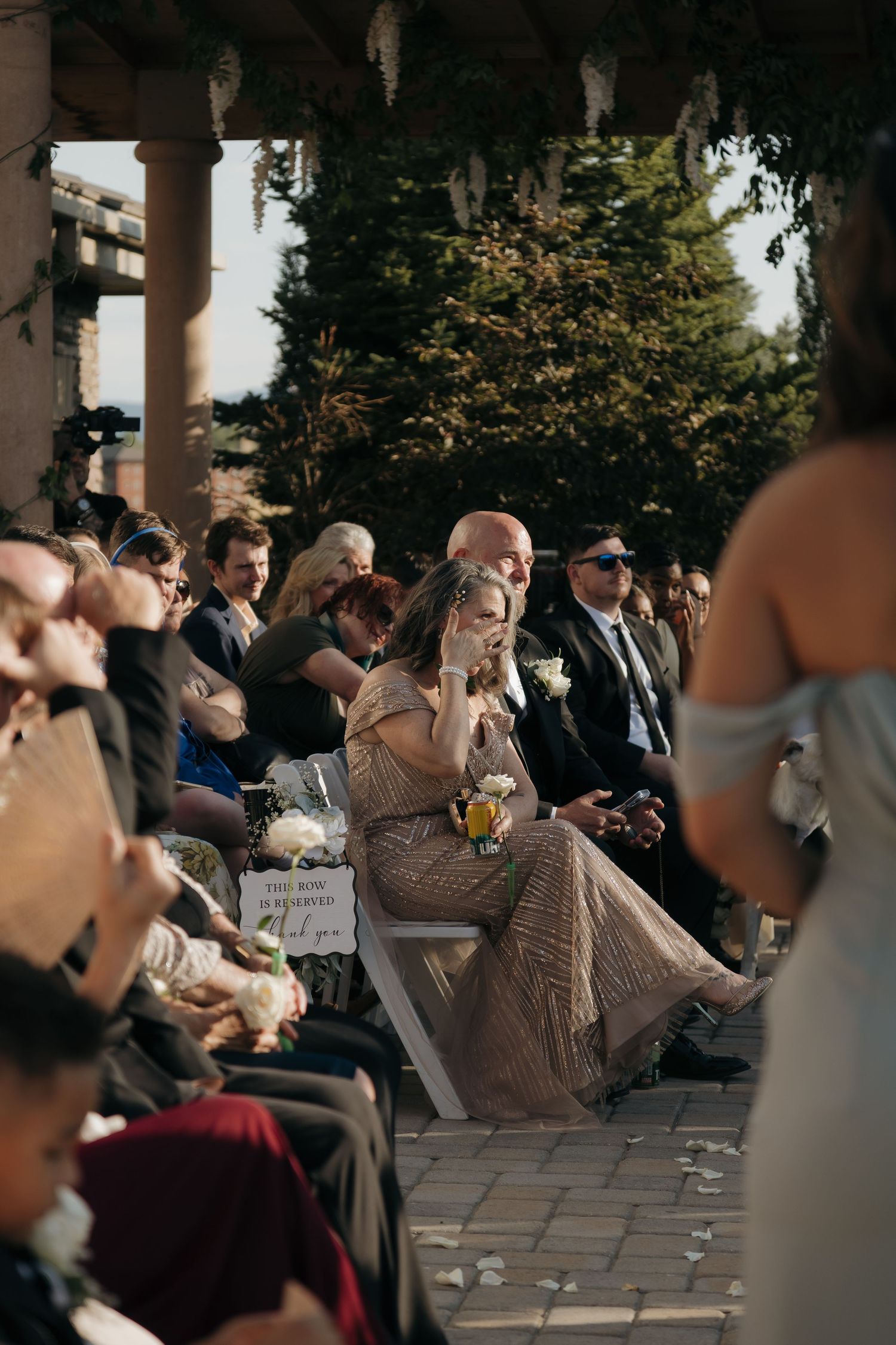 Tearful wedding guest in gold dress watching outdoor ceremony from decorated aisle seating.