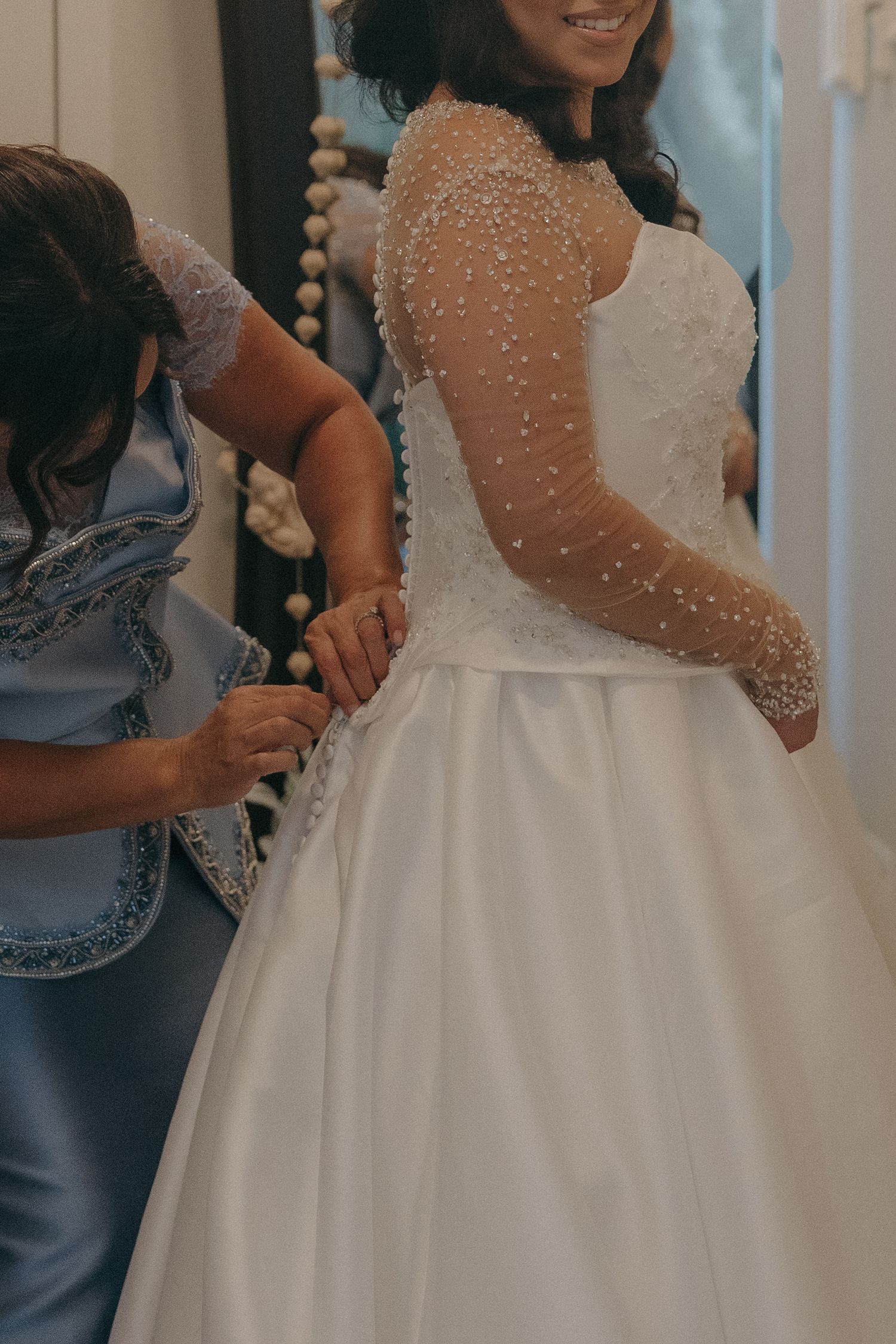 Bride in pearl-embellished wedding gown getting dress fastened by bridesmaid in blue dress.