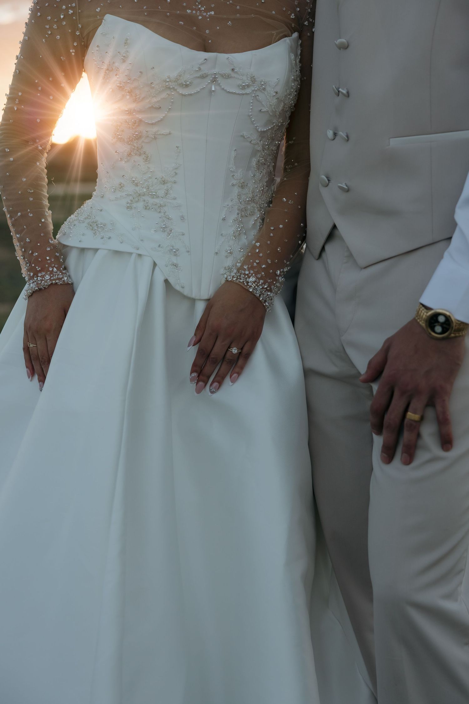 Close-up of bride in embroidered corset wedding gown and groom in ivory suit holding hands with wedding rings visible at sunset.