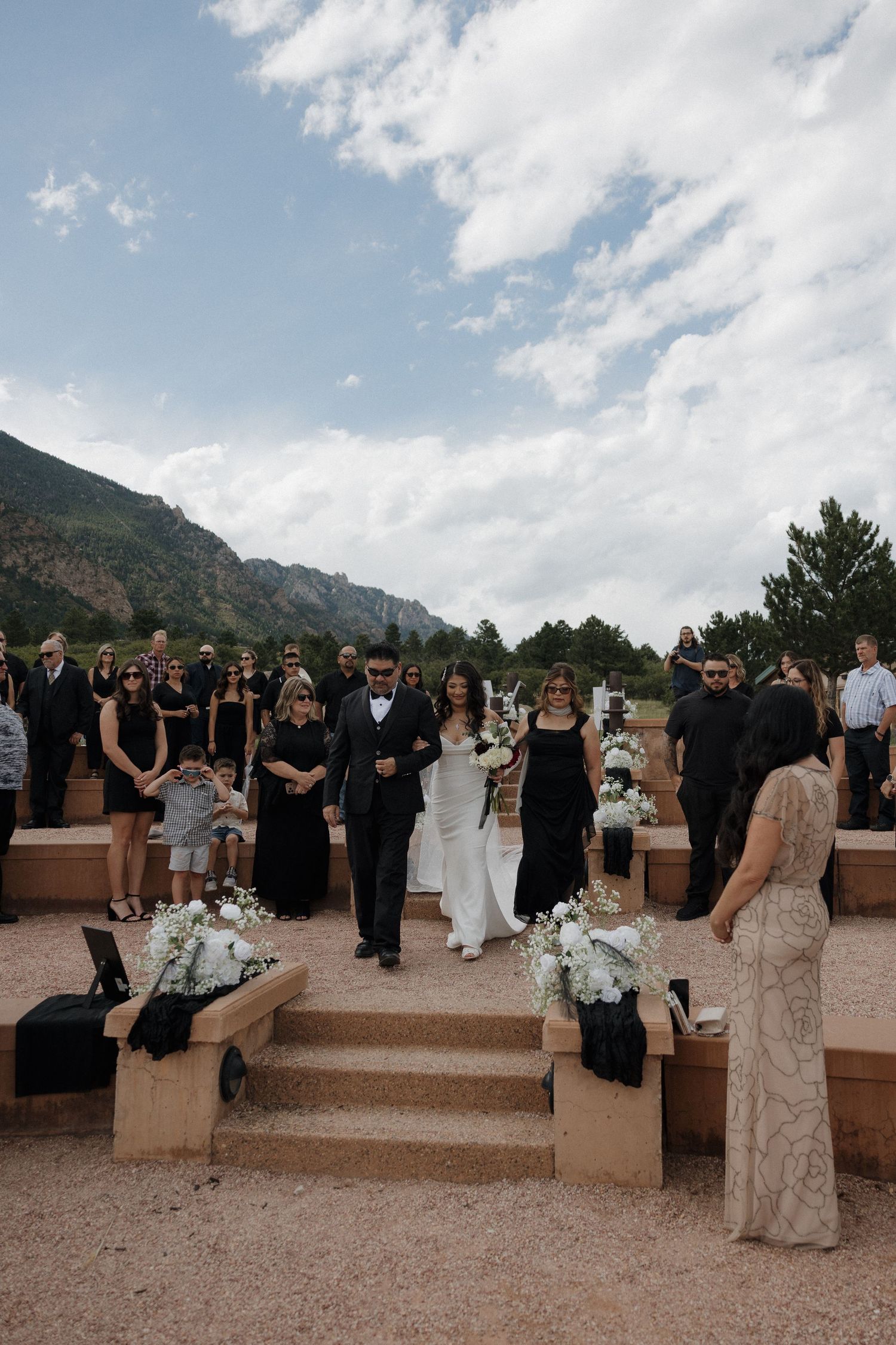 Bride walks down outdoor aisle with mountain backdrop during elegant black and white wedding ceremony.