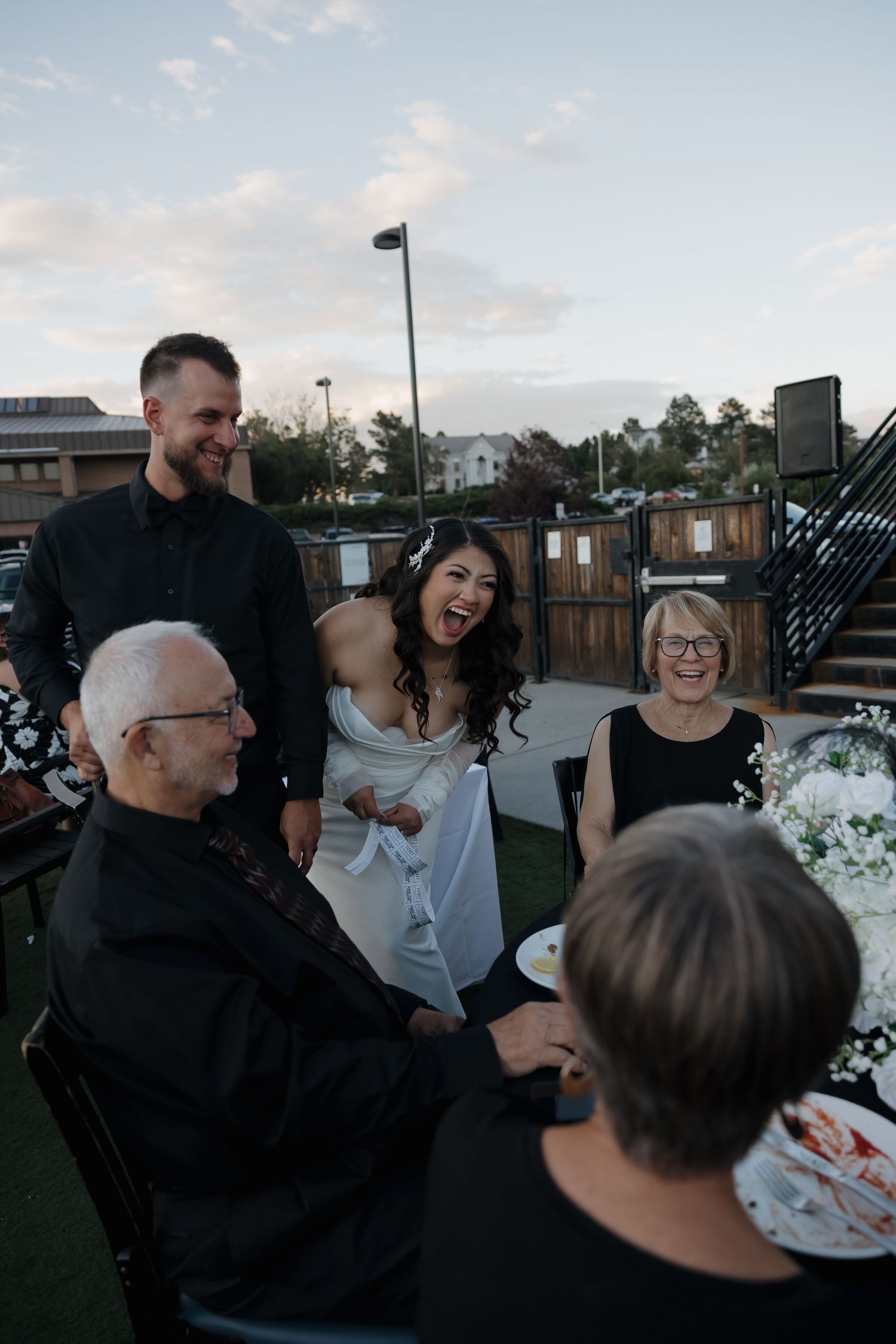 Bride and groom laughing with elderly guests at outdoor wedding reception dinner table at dusk.