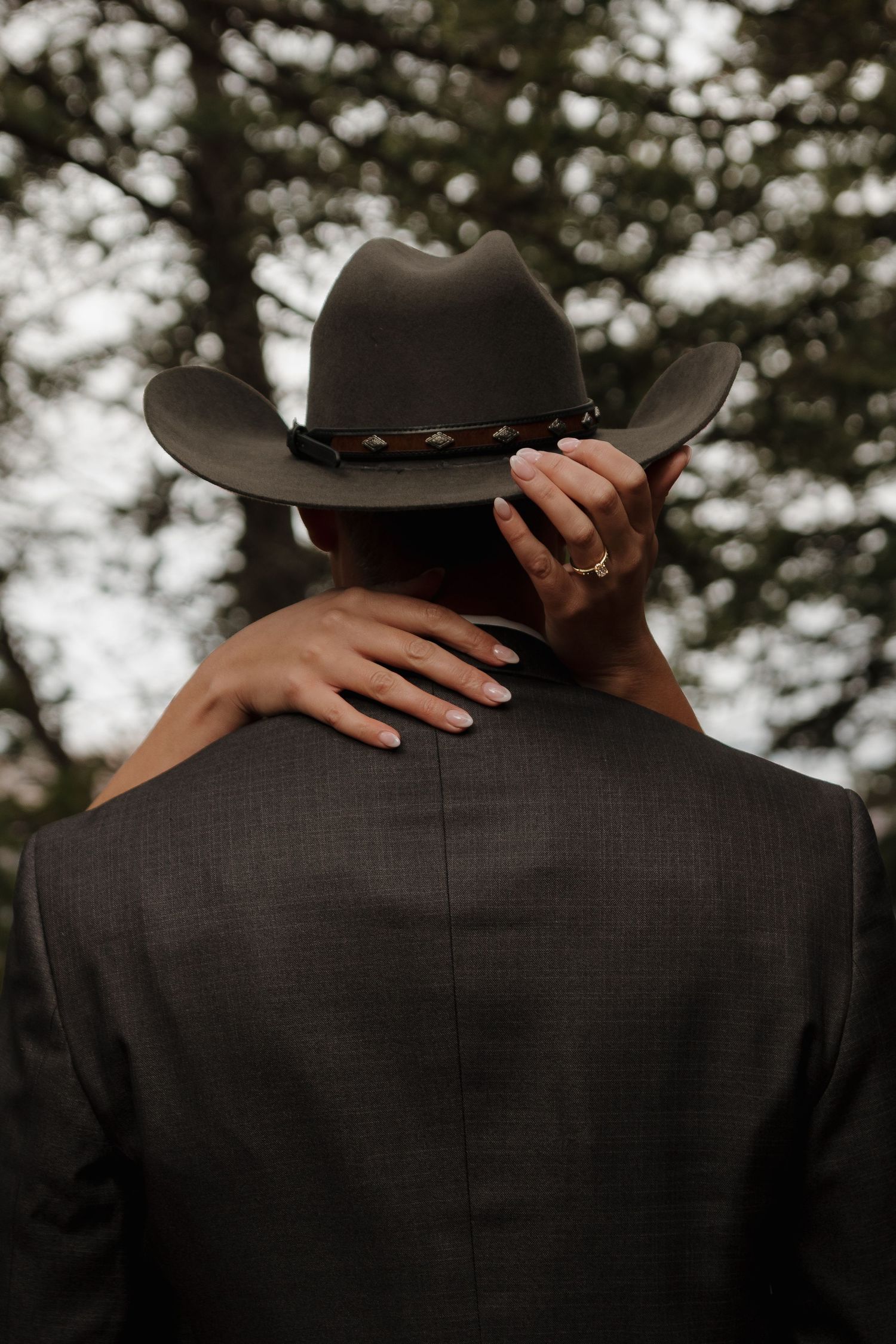 Back view of groom in dark suit and cowboy hat with bride's hands around his shoulders.