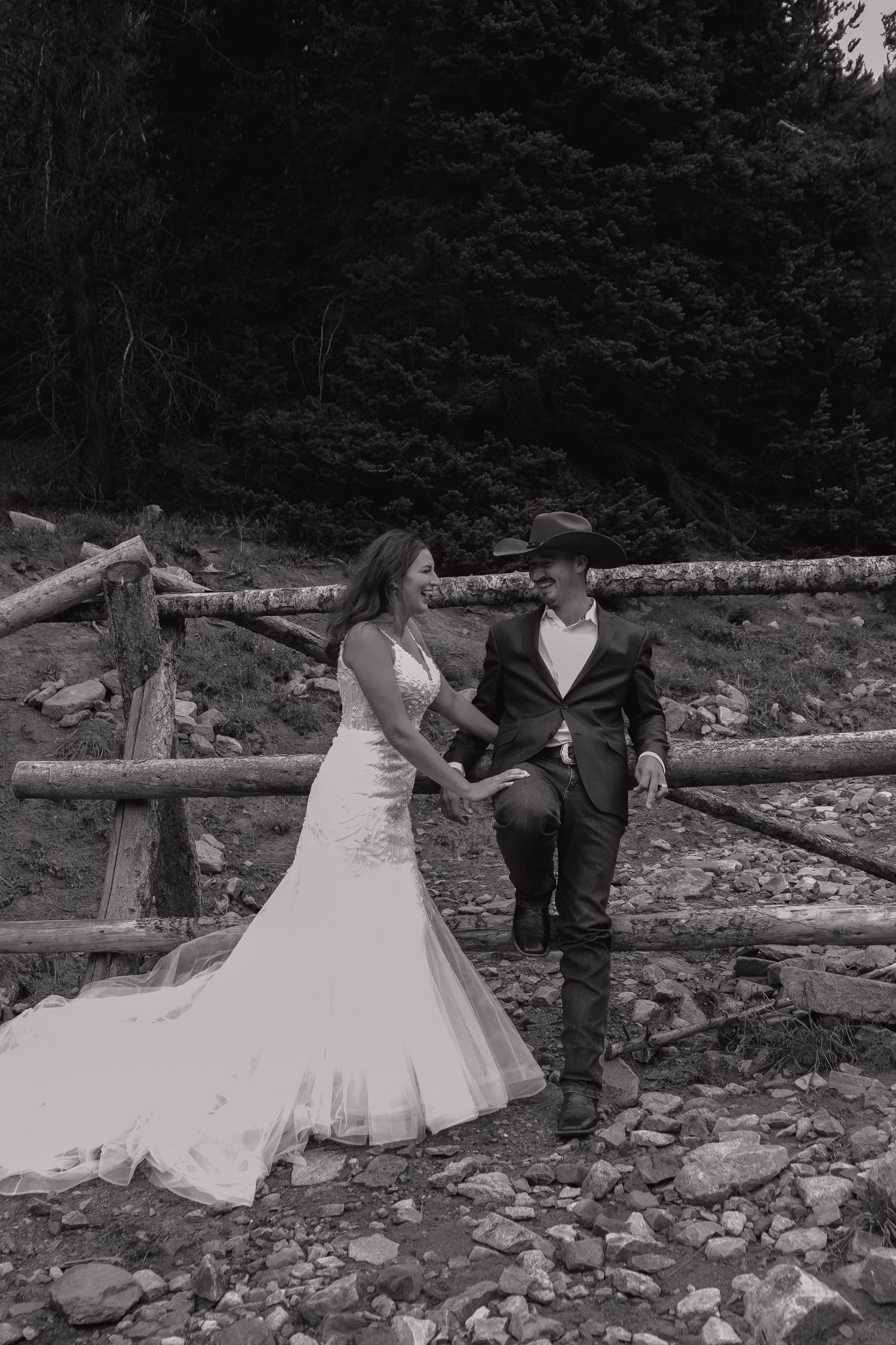 A couple in wedding attire poses on rocky terrain with a rustic wooden fence in the background.