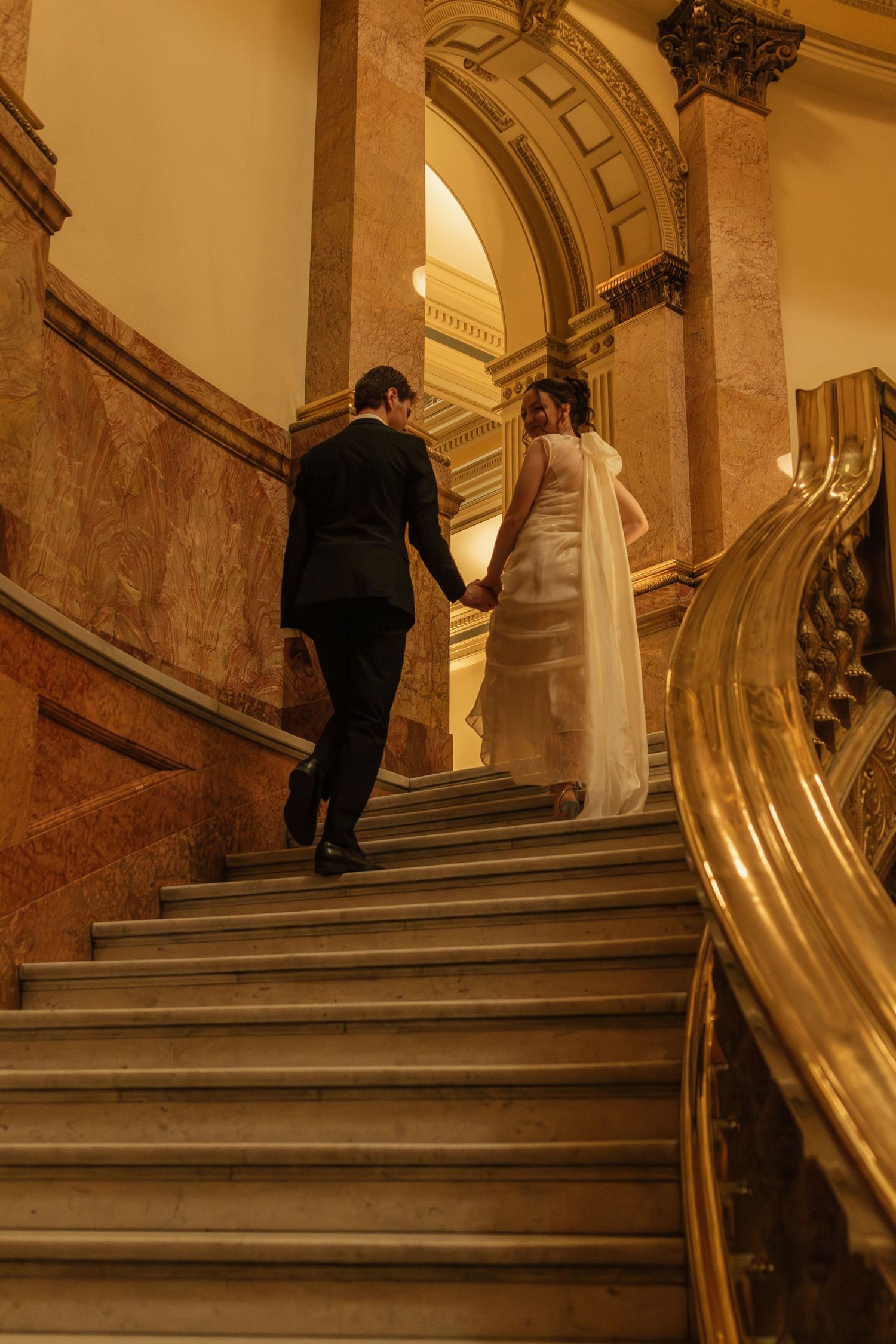 Couple ascending ornate marble stairs in formal wedding attire.