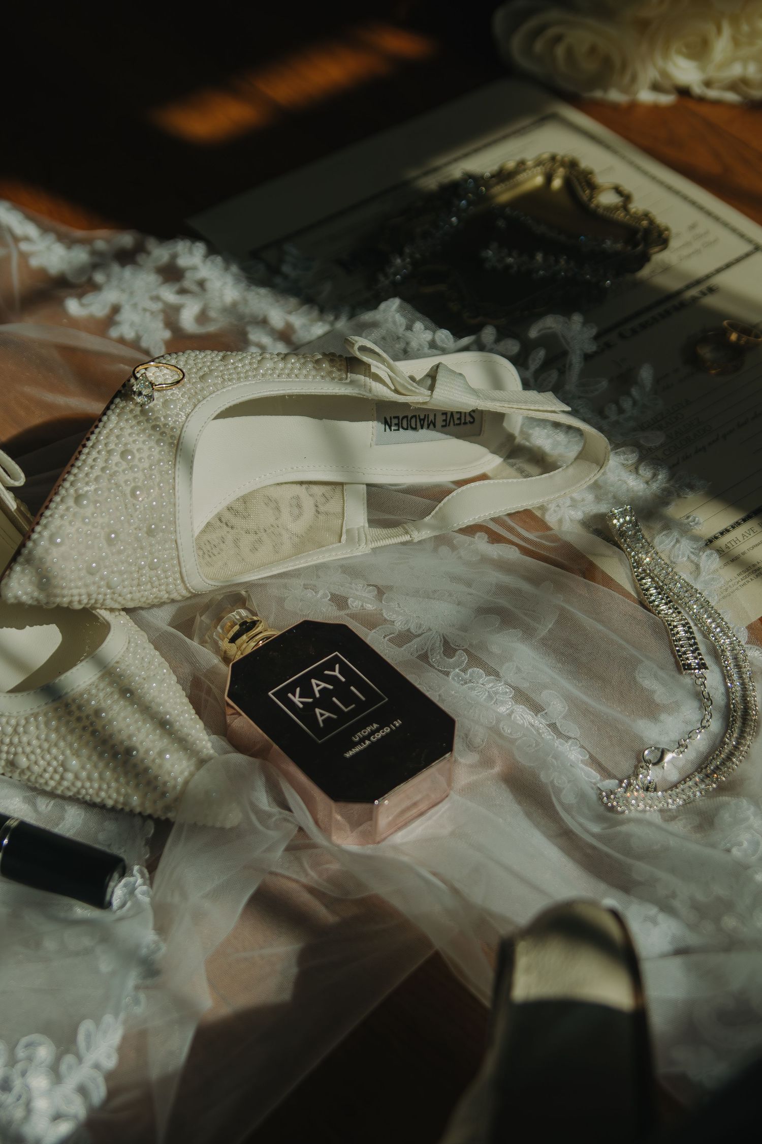 Wedding accessories including pearl necklaces, veil, and black perfume bottle arranged on a dark surface in moody lighting.
