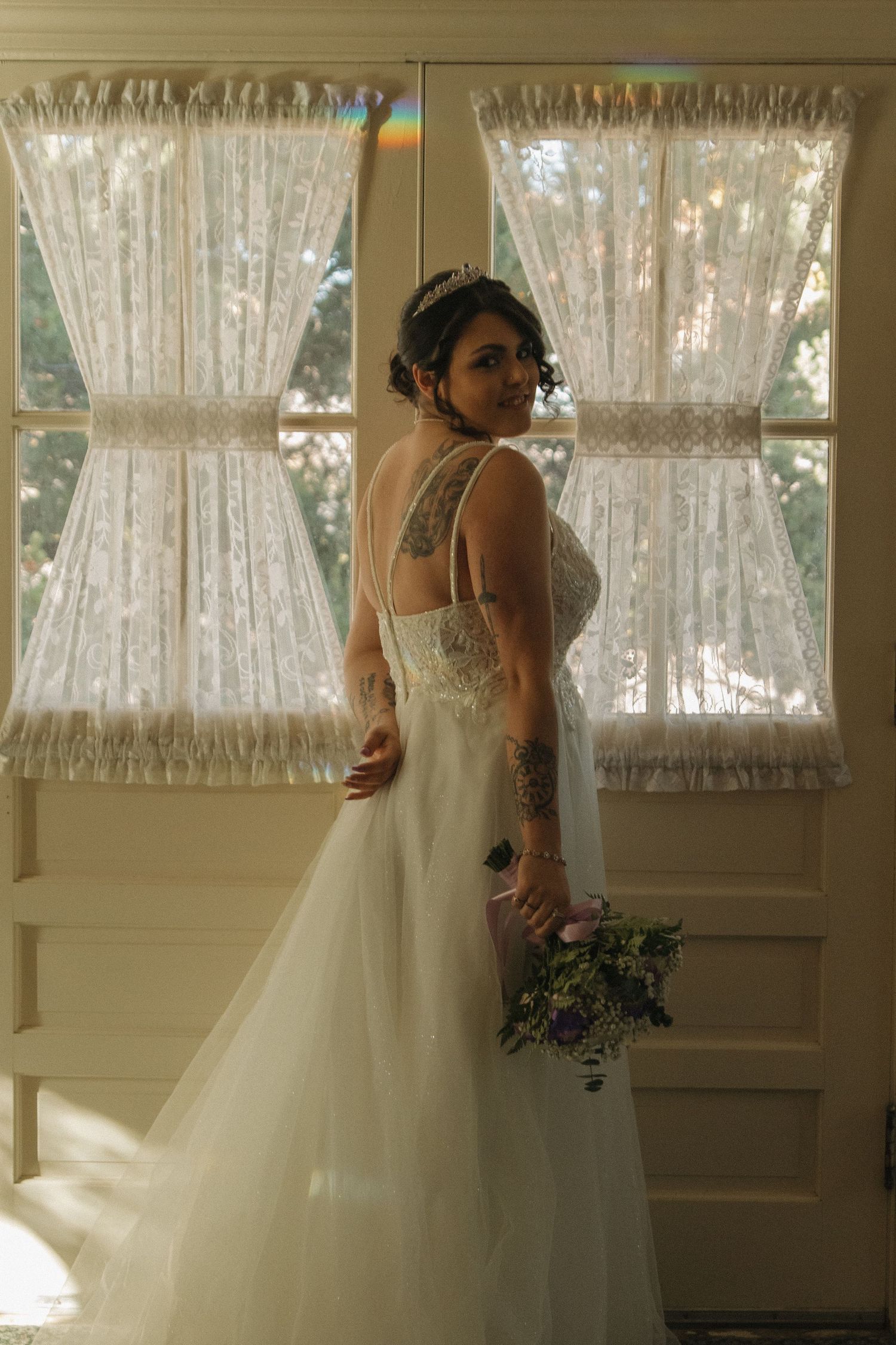A bride in a flowing white tulle gown stands by sunlit windows in a vintage-style room.
