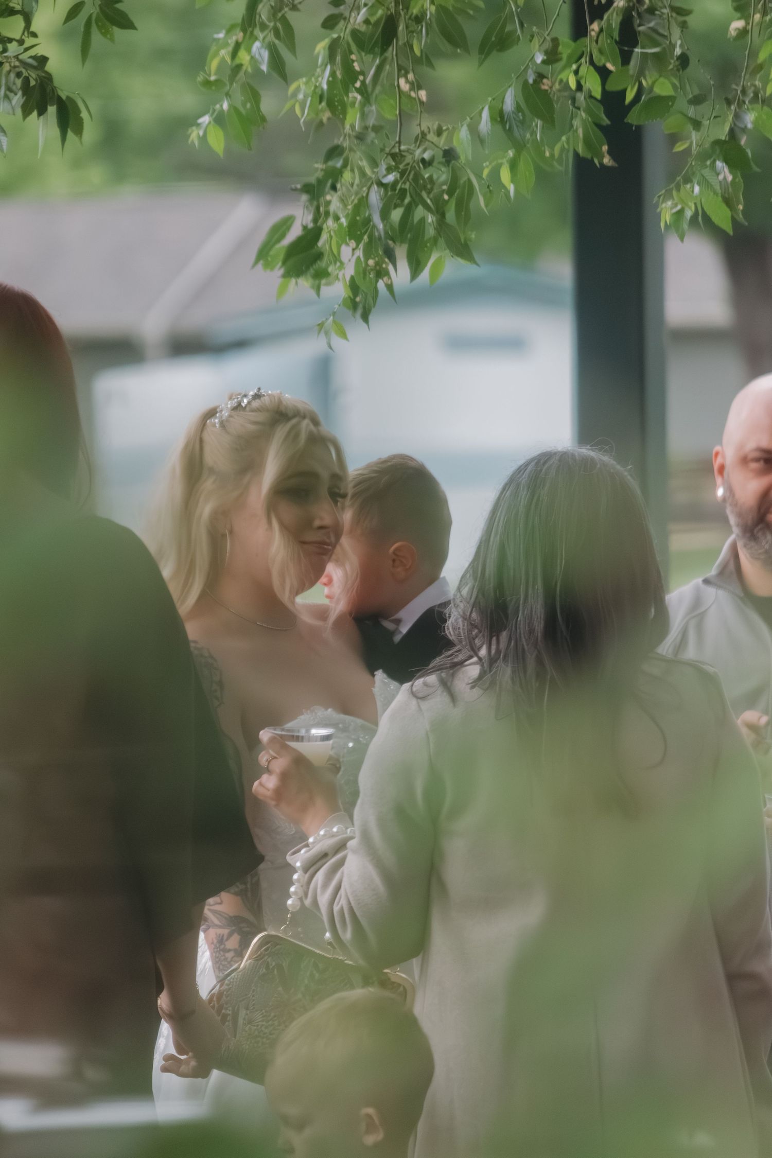 Wedding guests gather under green foliage at an outdoor celebration.
