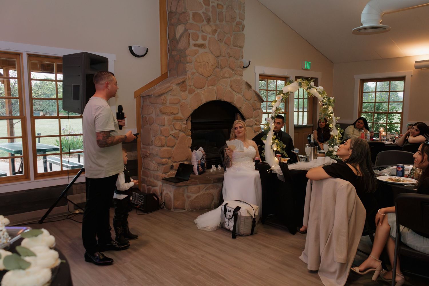 Wedding reception taking place in a rustic venue with stone fireplace and hardwood floors during speeches.