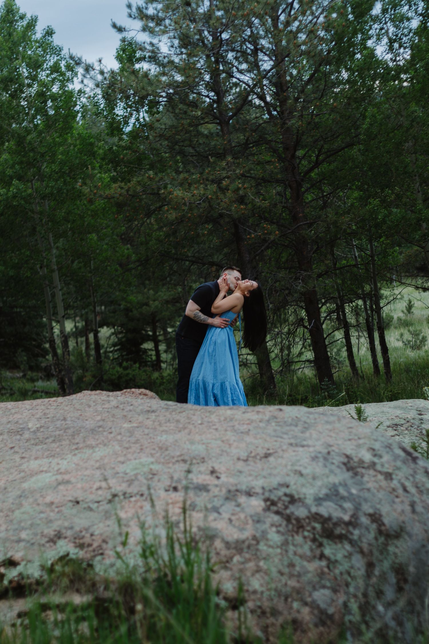 A couple in formal attire sharing a romantic moment on a boulder in a forest setting.