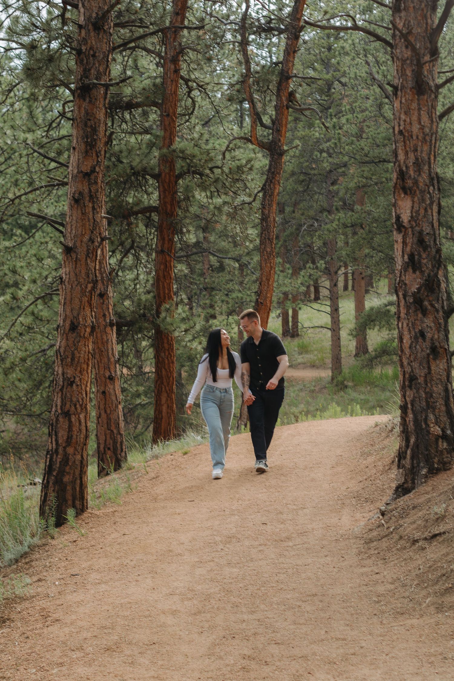 A couple walks together on a dirt path through towering pine trees in a mountain forest.