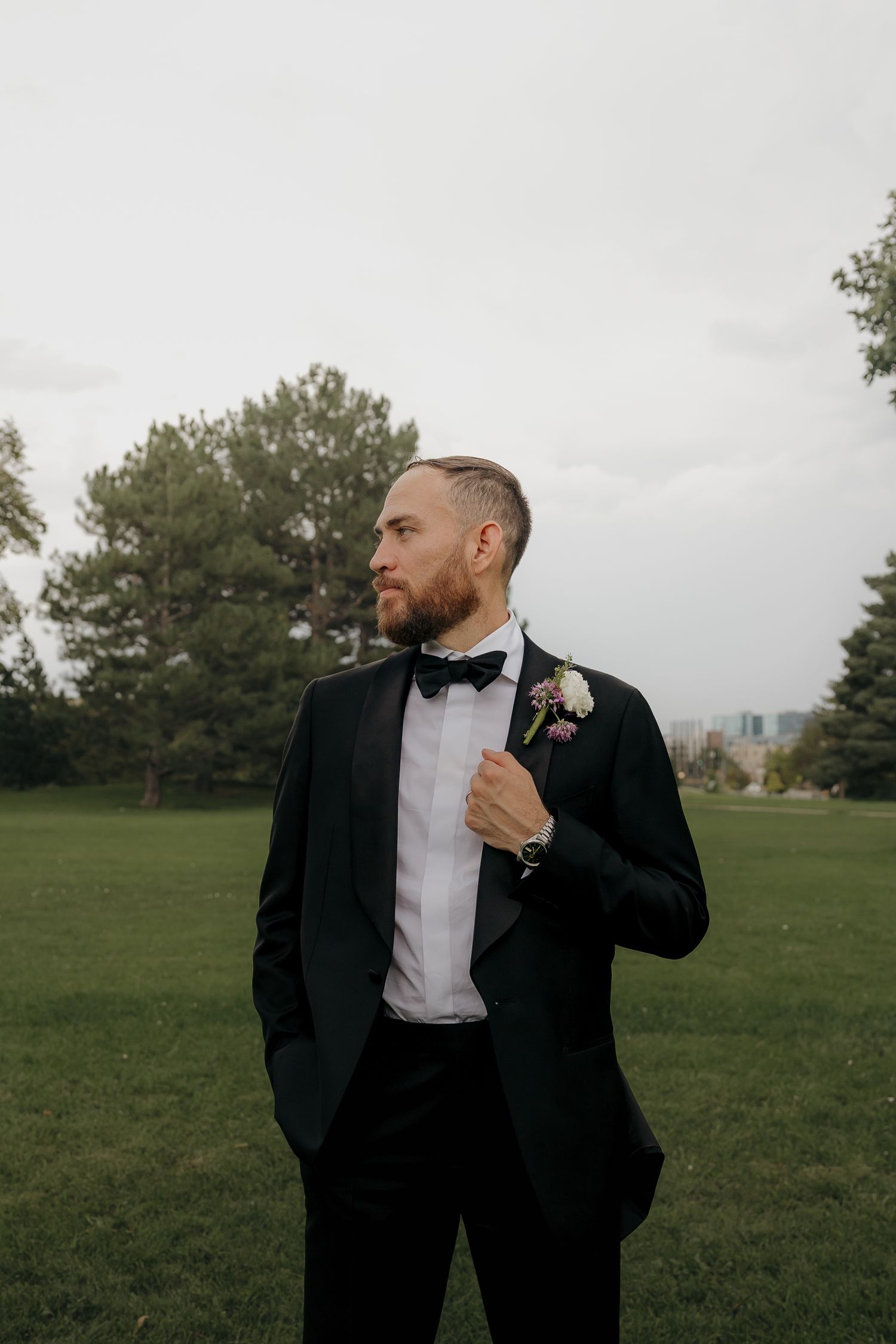 Dapper groom in black tuxedo with boutonniere standing confidently in green park setting.
