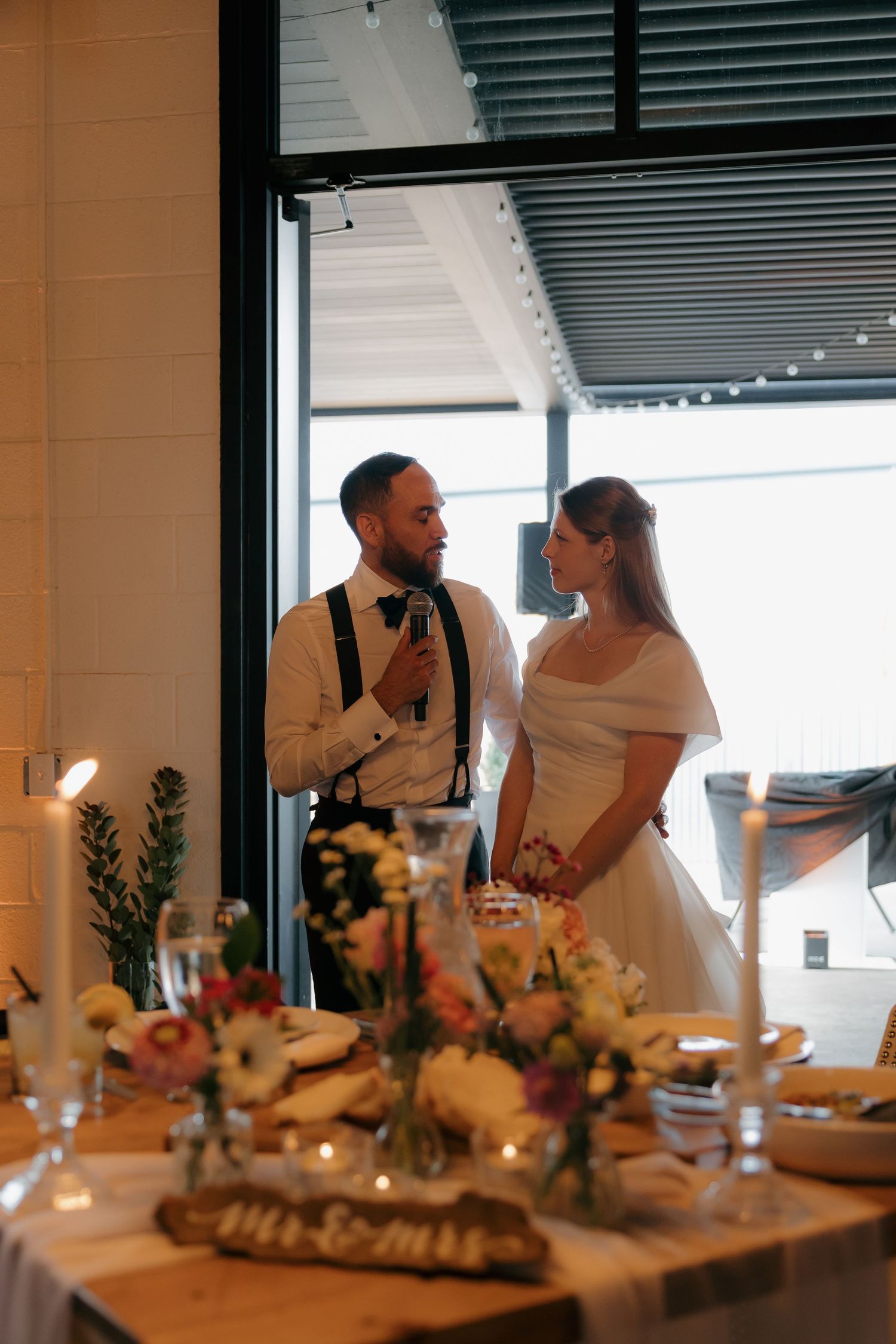 Newlywed couple in white gown and suspenders share intimate moment beside elegantly decorated reception dinner table.