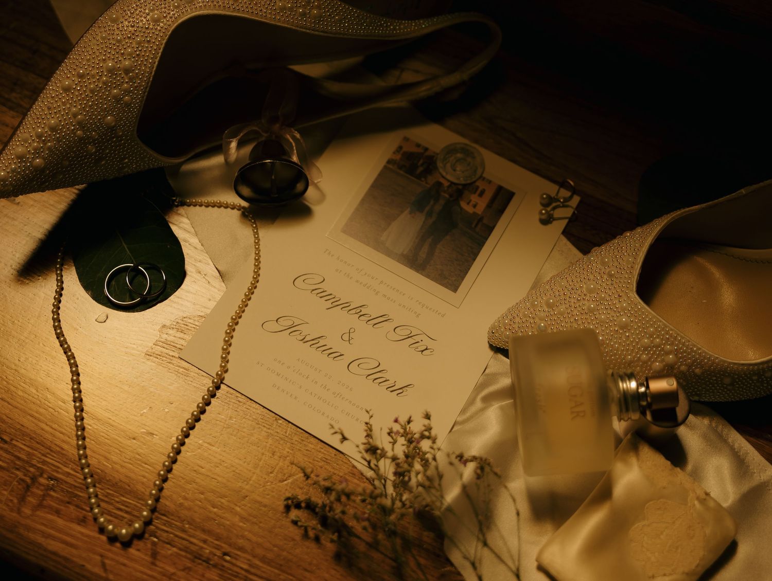 Wedding invitation with couple photo, pearl necklace, ring, perfume bottle, and glittery heels on wooden surface.