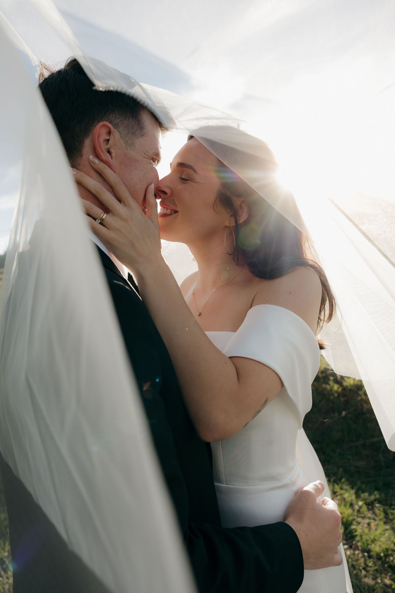 Bride and groom sharing romantic almost-kiss under veil in golden sunlight.