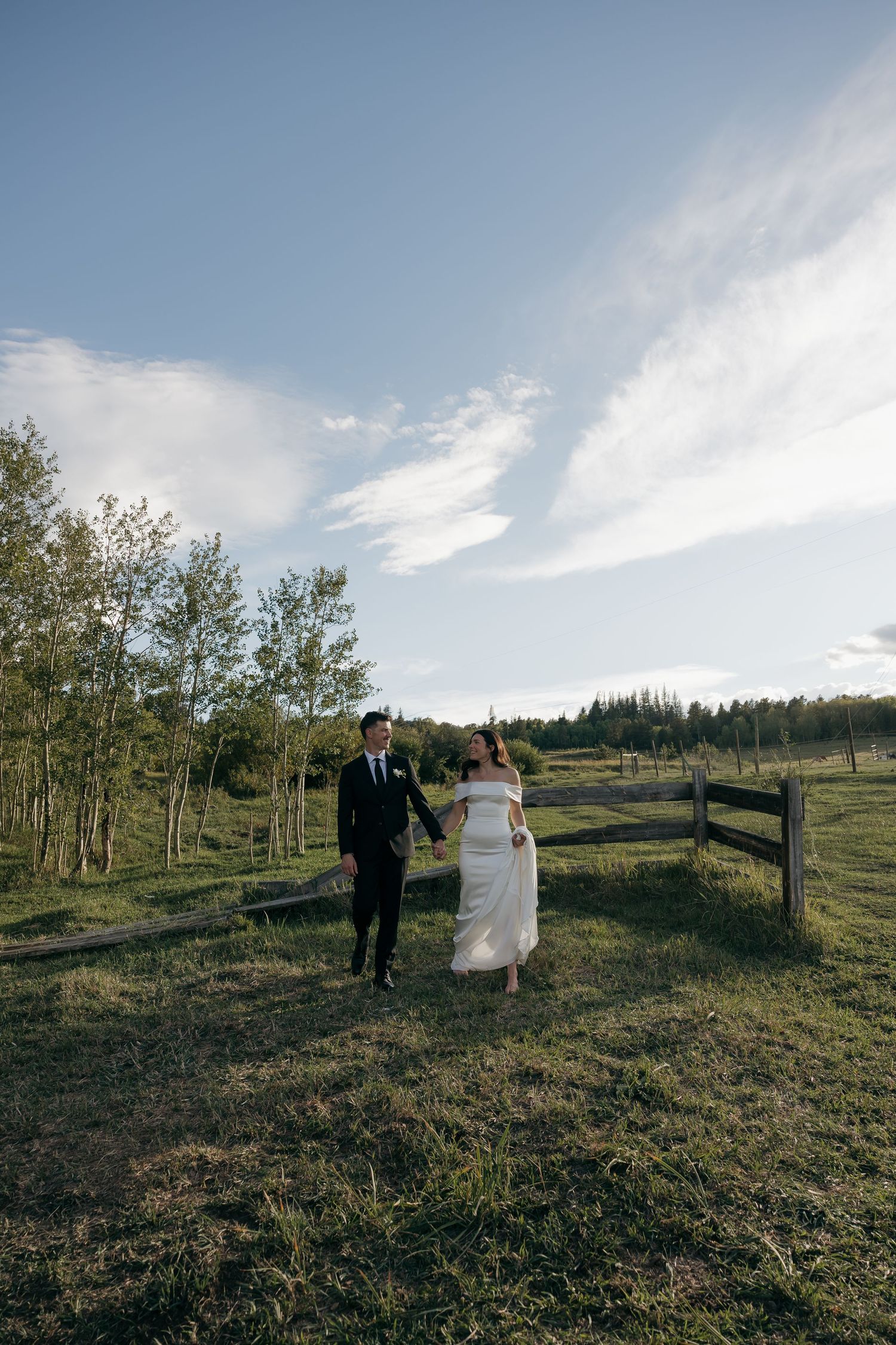 Bride and groom walking hand in hand through a scenic green pasture with wooden fence and blue sky backdrop.