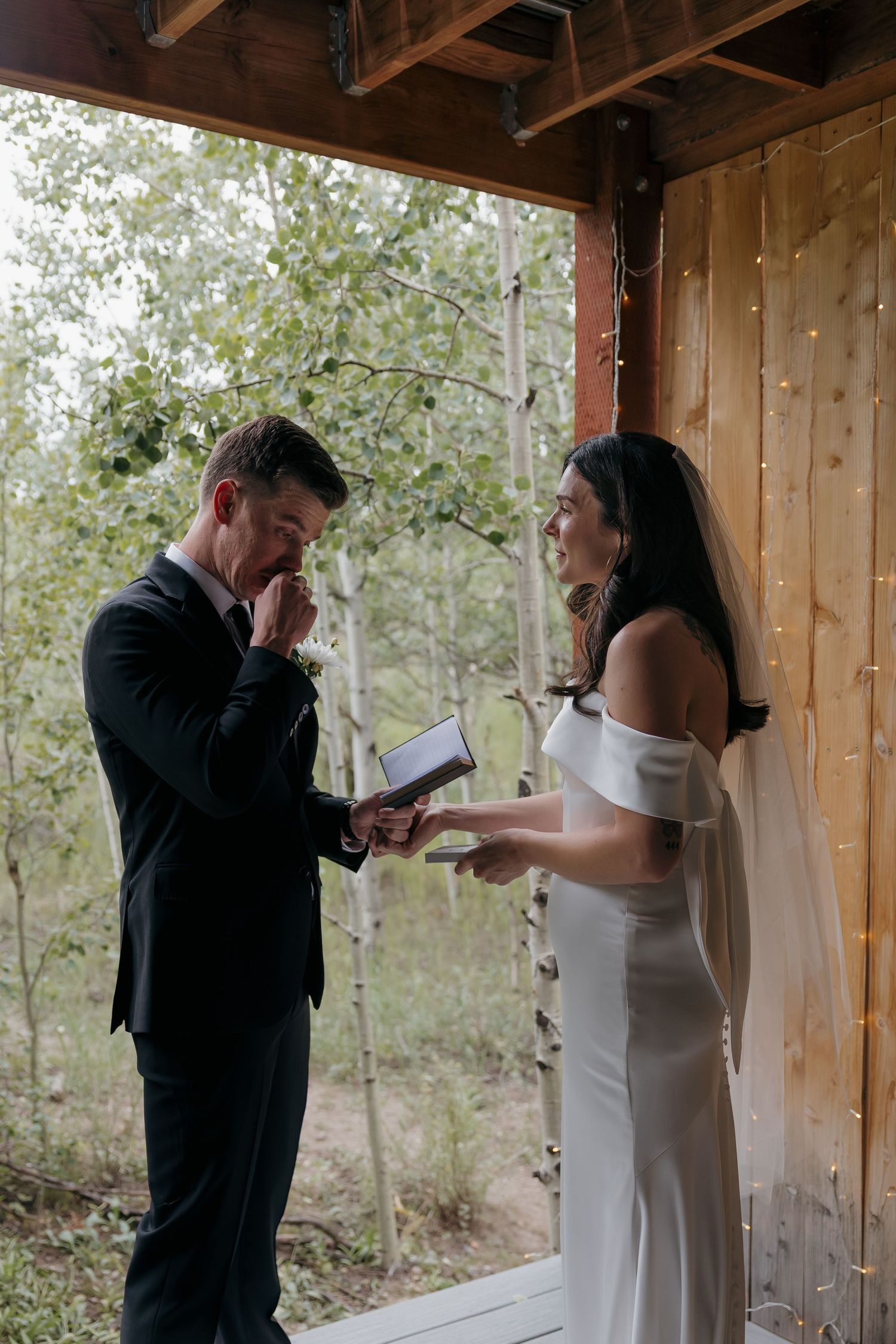 Emotional groom reading vows to bride in white dress and veil during outdoor wedding ceremony on rustic wooden porch.