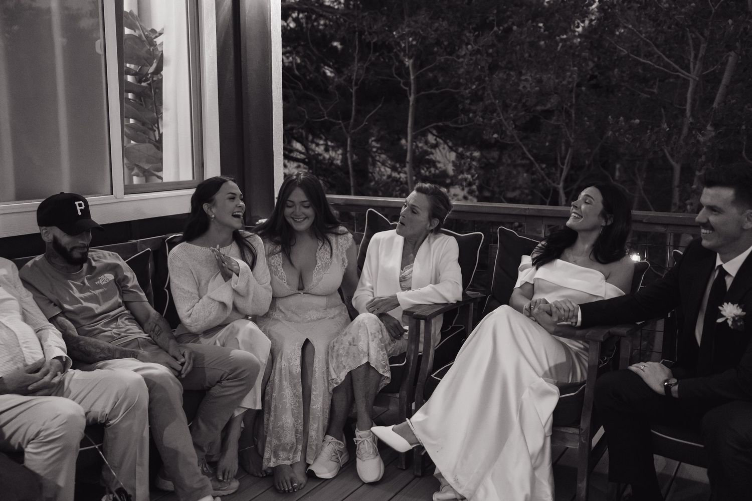 Black and white candid photo of bride, groom, and wedding guests laughing together on an outdoor patio at night.