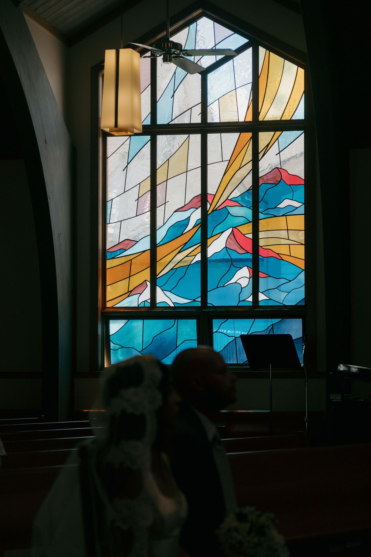 Bride and groom silhouetted before a colorful stained glass window inside a church during wedding ceremony.
