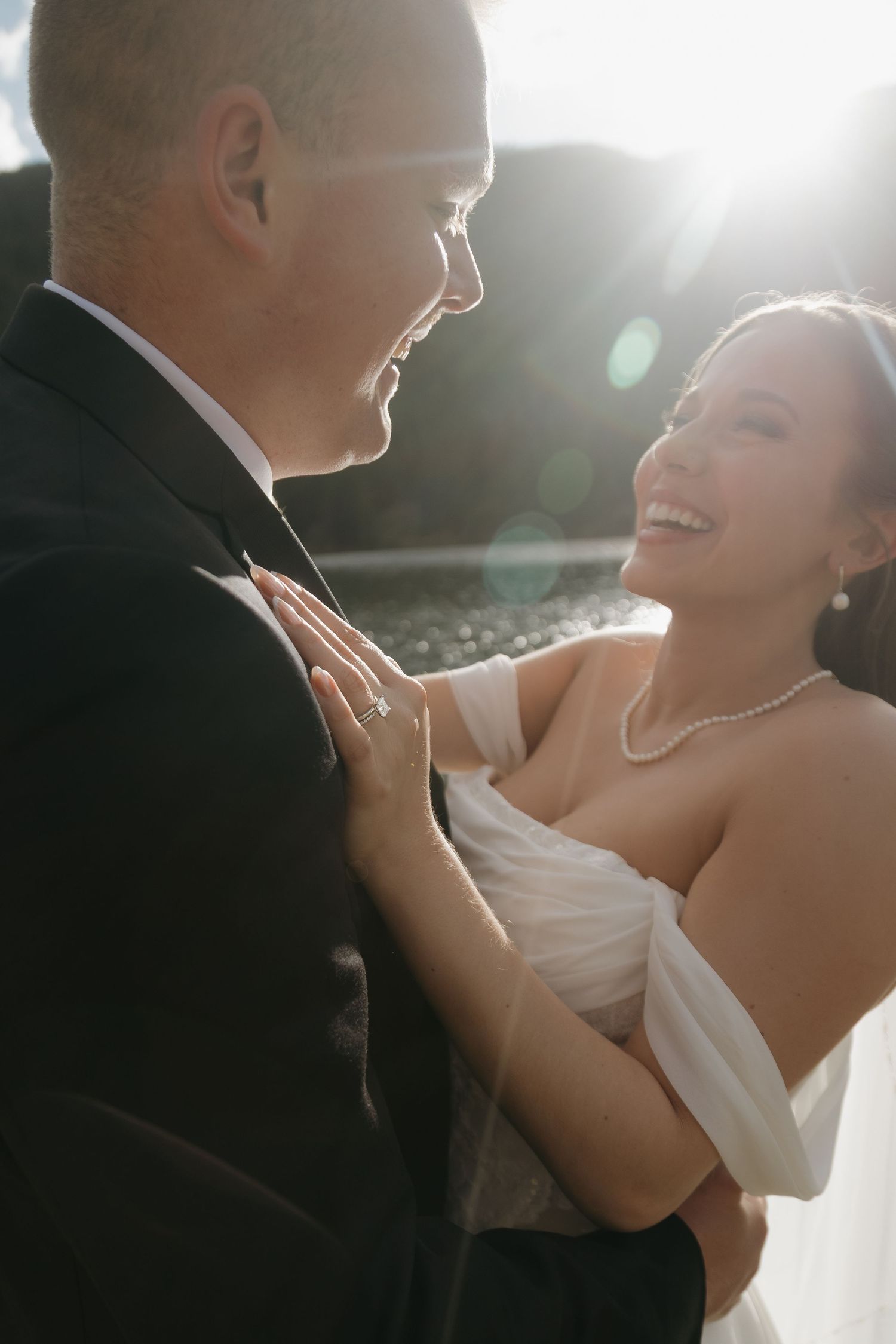 Laughing bride with pearl necklace embraces groom in a sunlit, joyful moment during their outdoor wedding.