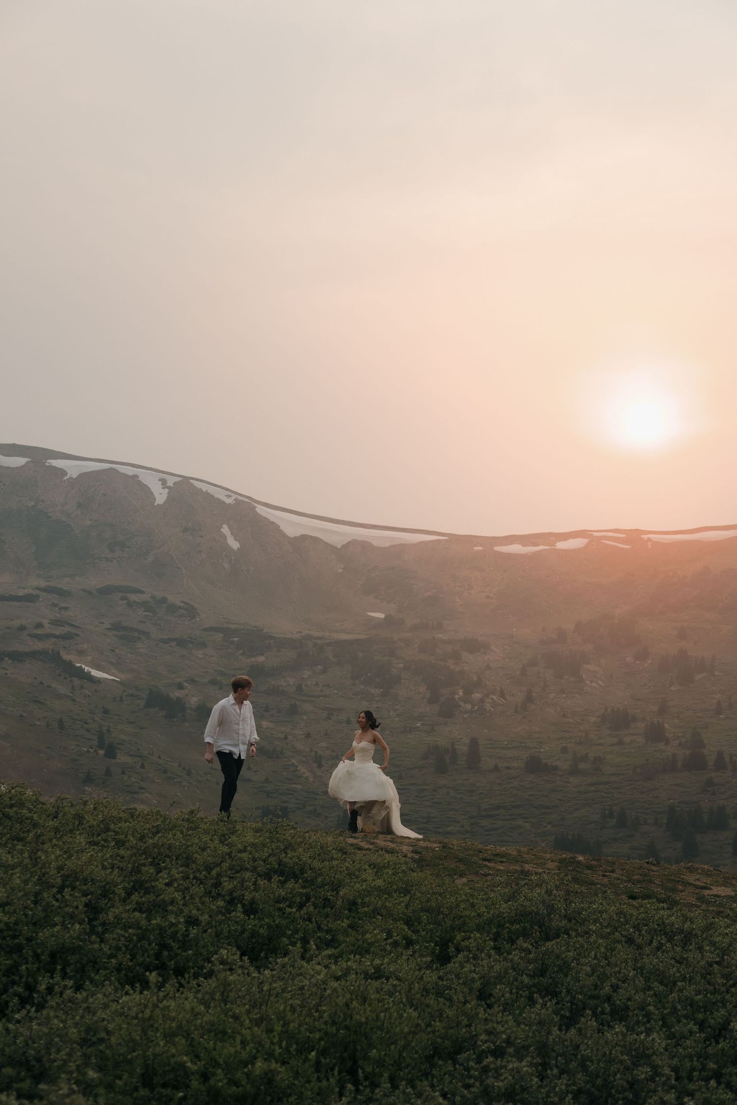 Bride and groom walking on misty mountain hillside at sunset during outdoor wedding photoshoot.
