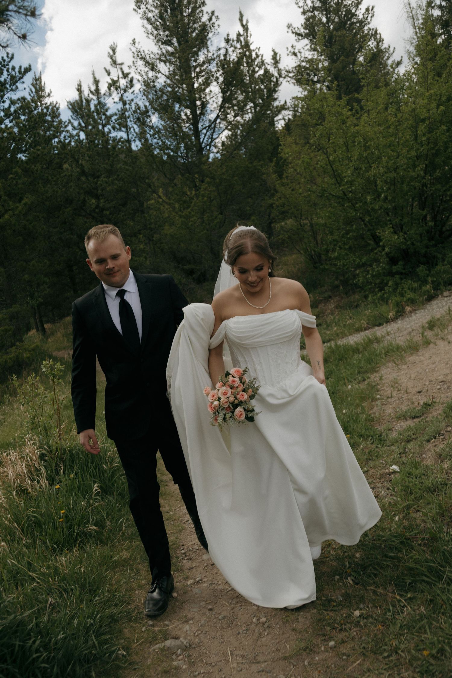 Smiling bride and groom walk hand-in-hand along a forest trail during their outdoor mountain wedding celebration.