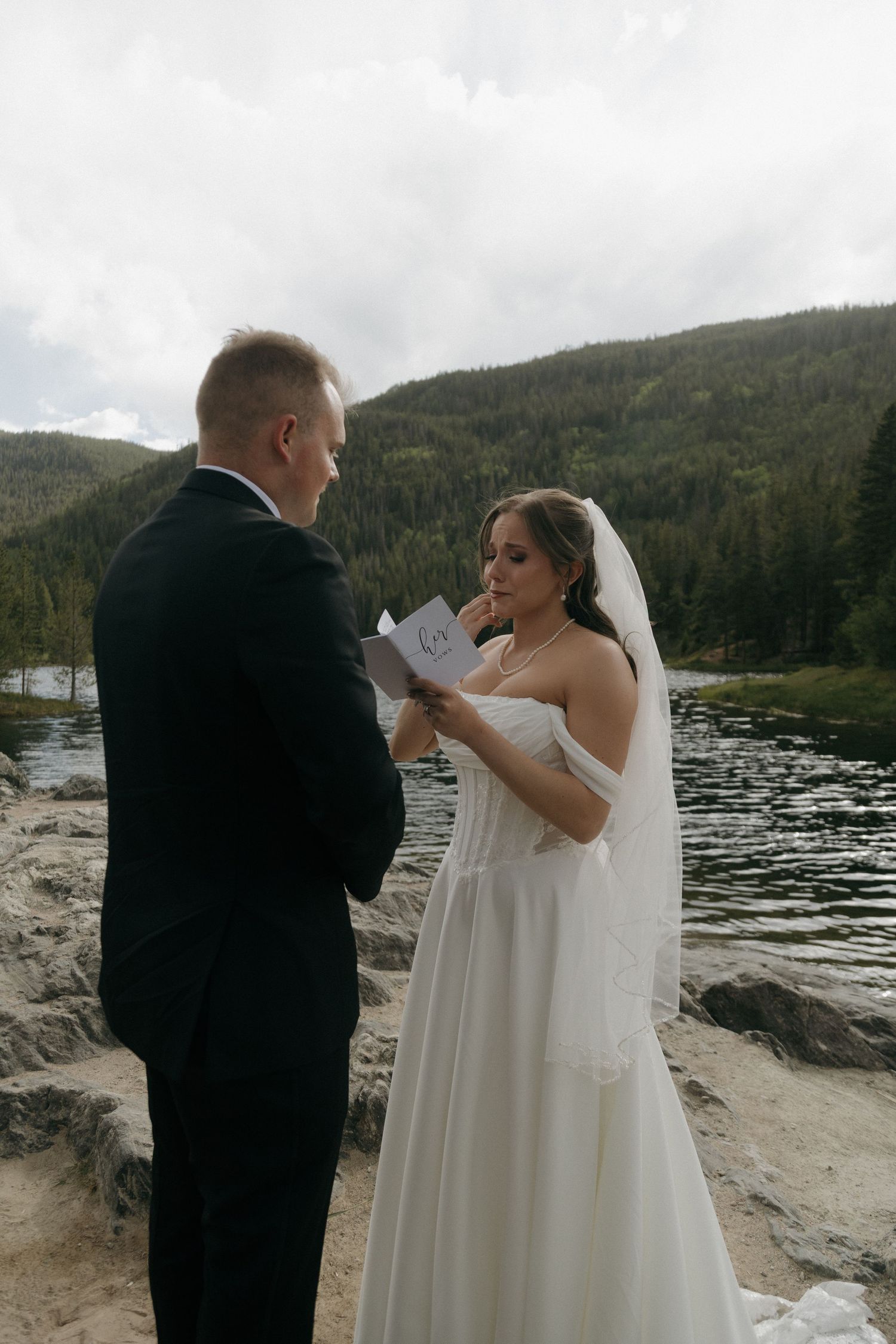 Bride reads wedding vows from a card while groom listens near a mountain lake during outdoor elopement ceremony.