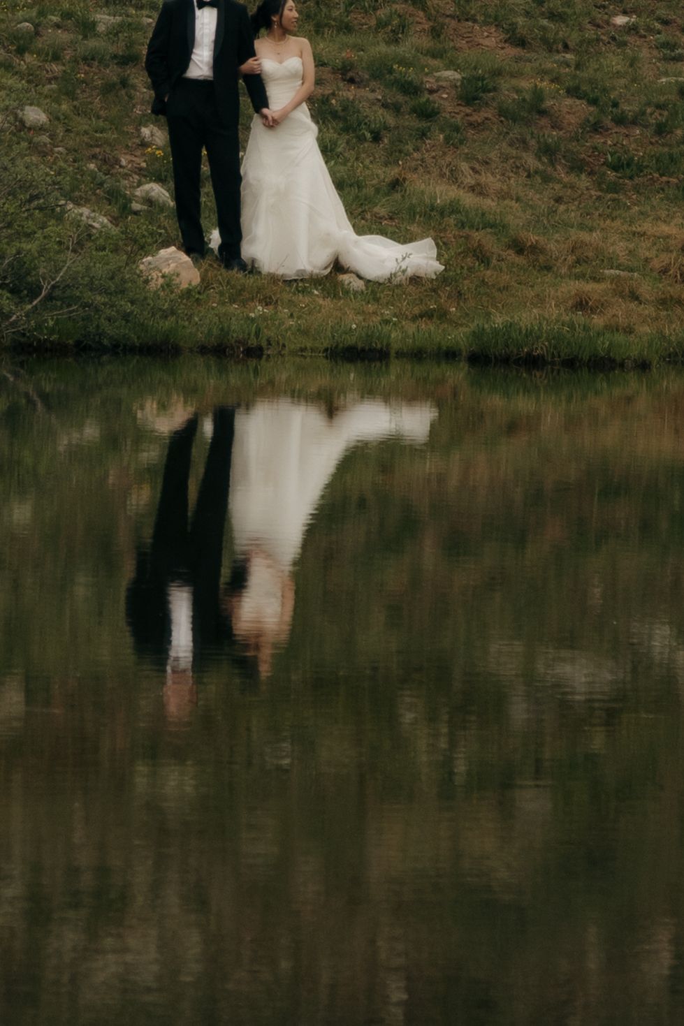 A bride and groom stand by a reflective mountain lake, their mirror image visible in the still water below.