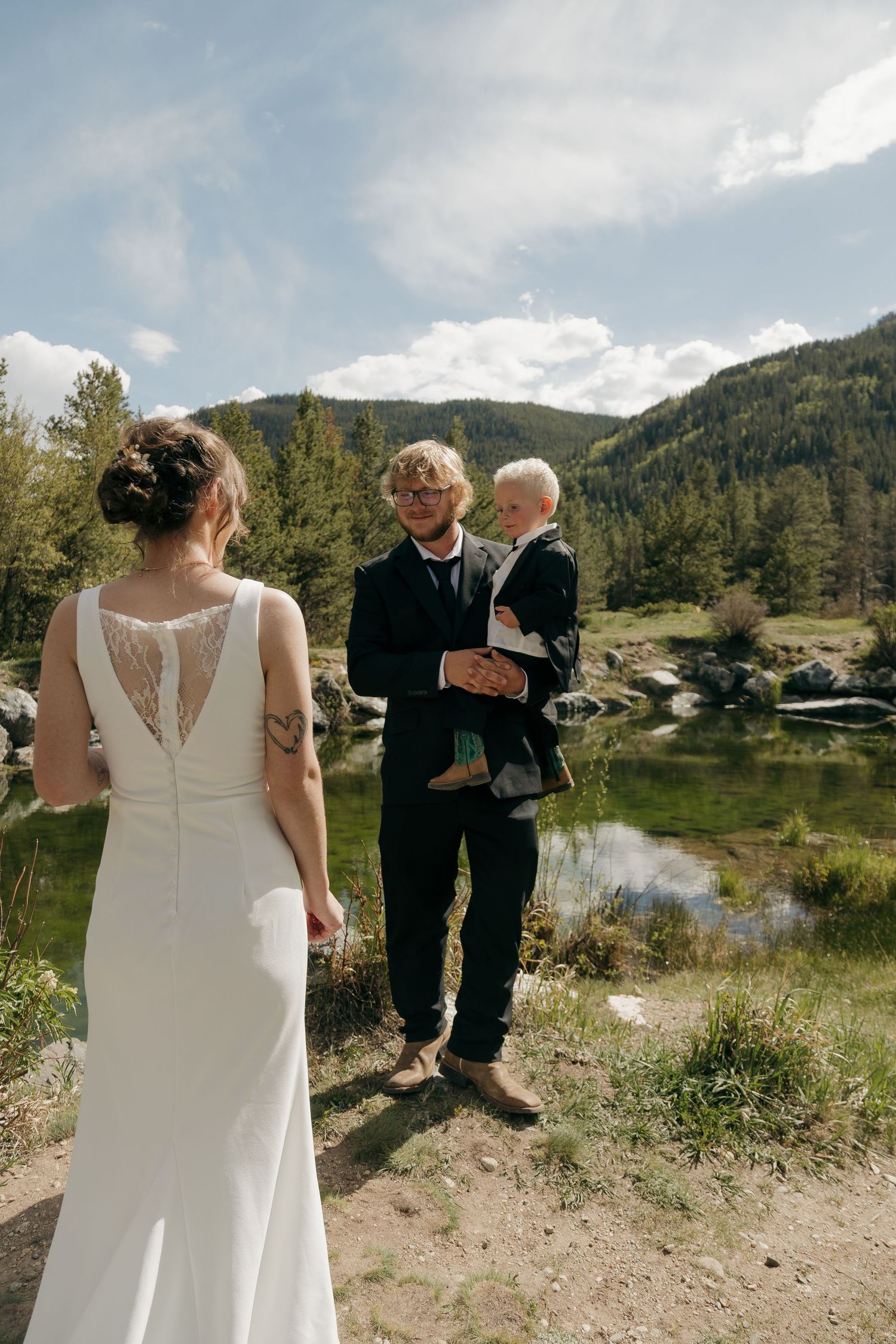 Bride in white lace-back gown faces groom holding baby boy in suit by mountain lake.